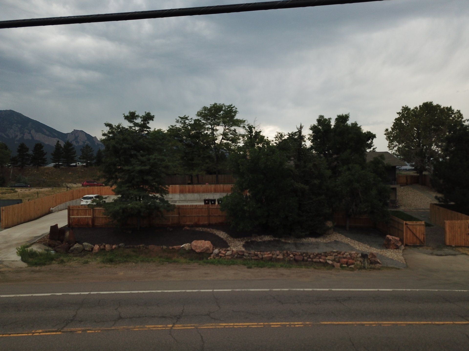 Roadside view of a fenced property with trees and a mountain backdrop under an overcast sky.