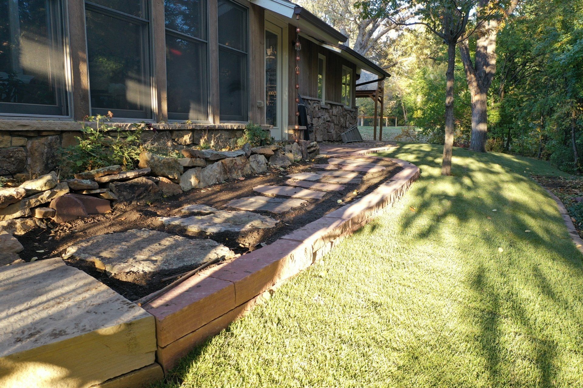 Stone path and retaining wall by a house and lawn, surrounded by trees.
