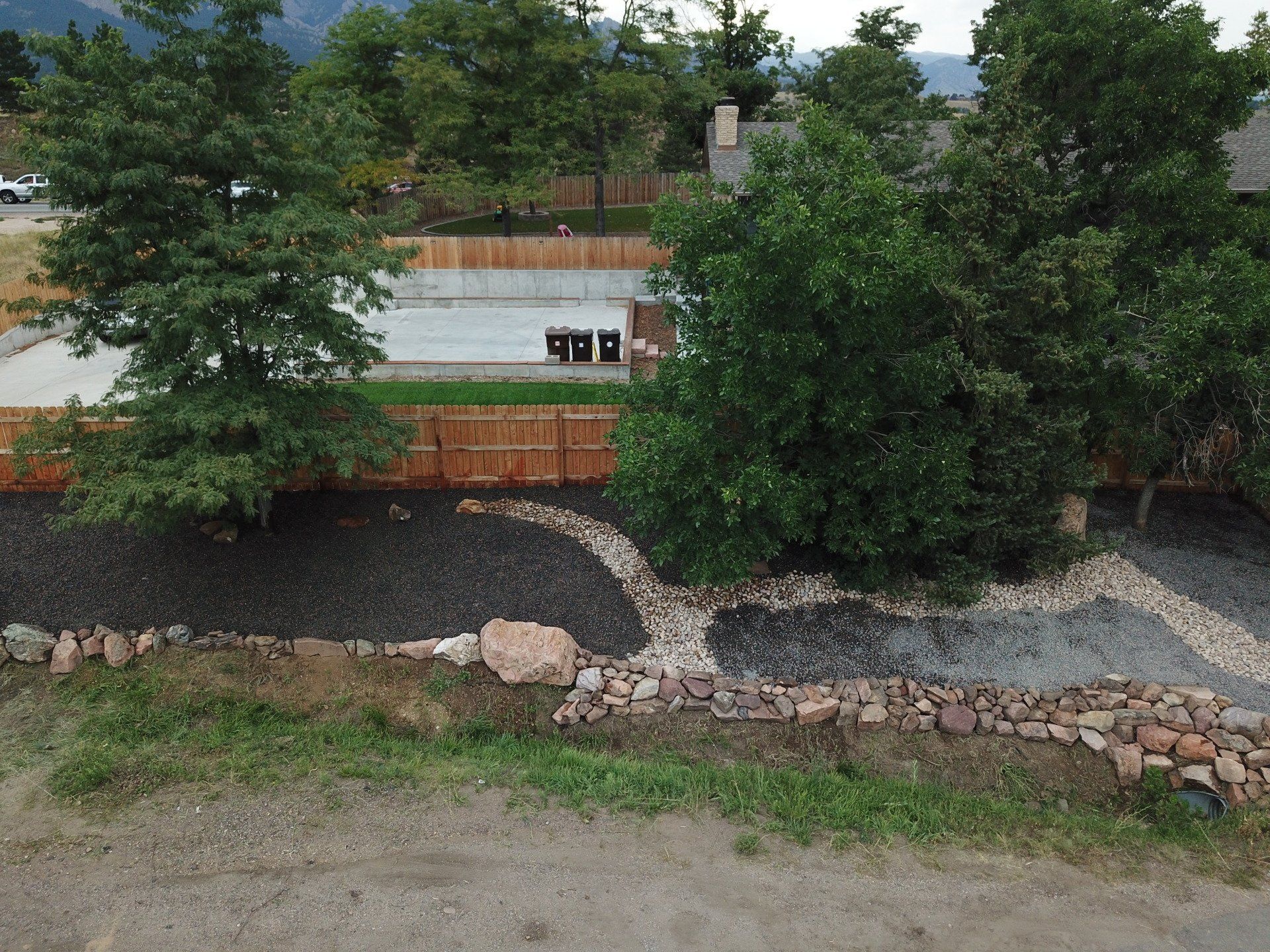 View of landscaping with trees, rocks, gravel, and a wooden fence. Mountains in the background.