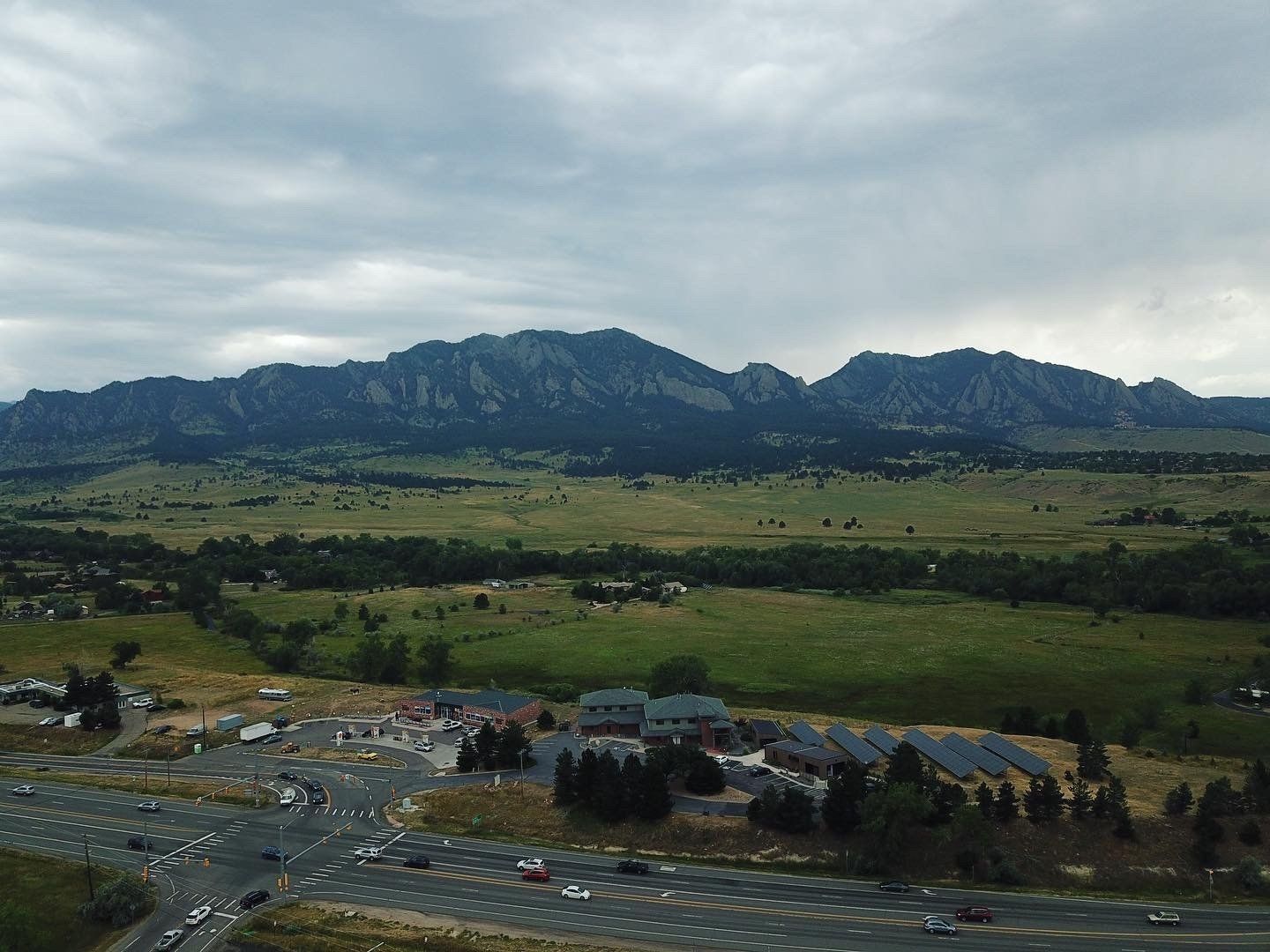 Mountain range over green fields, small town with busy roads, cloudy sky.
