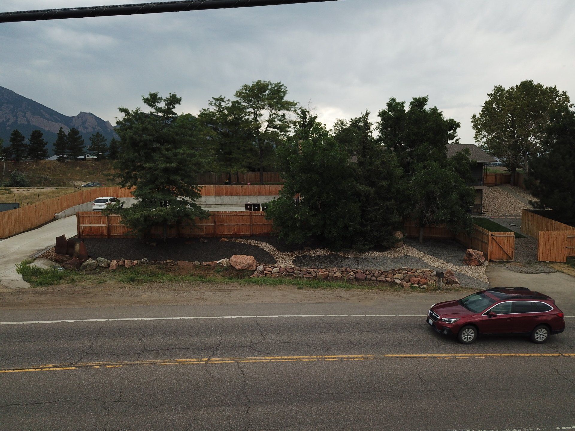 A brown car drives on a road; in the background, a fenced property with trees and a mountain under a cloudy sky.