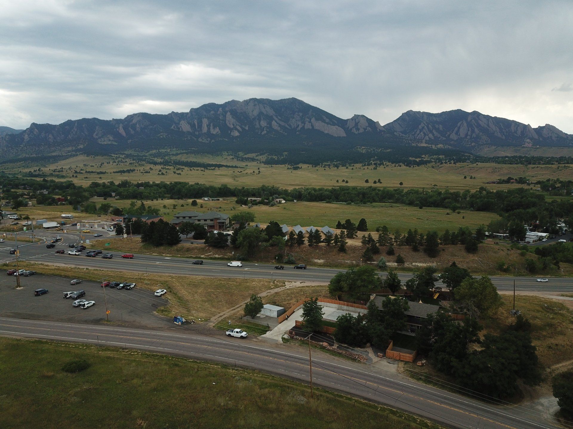 Mountains loom over a valley with buildings, roads, and a parking lot under a cloudy sky.