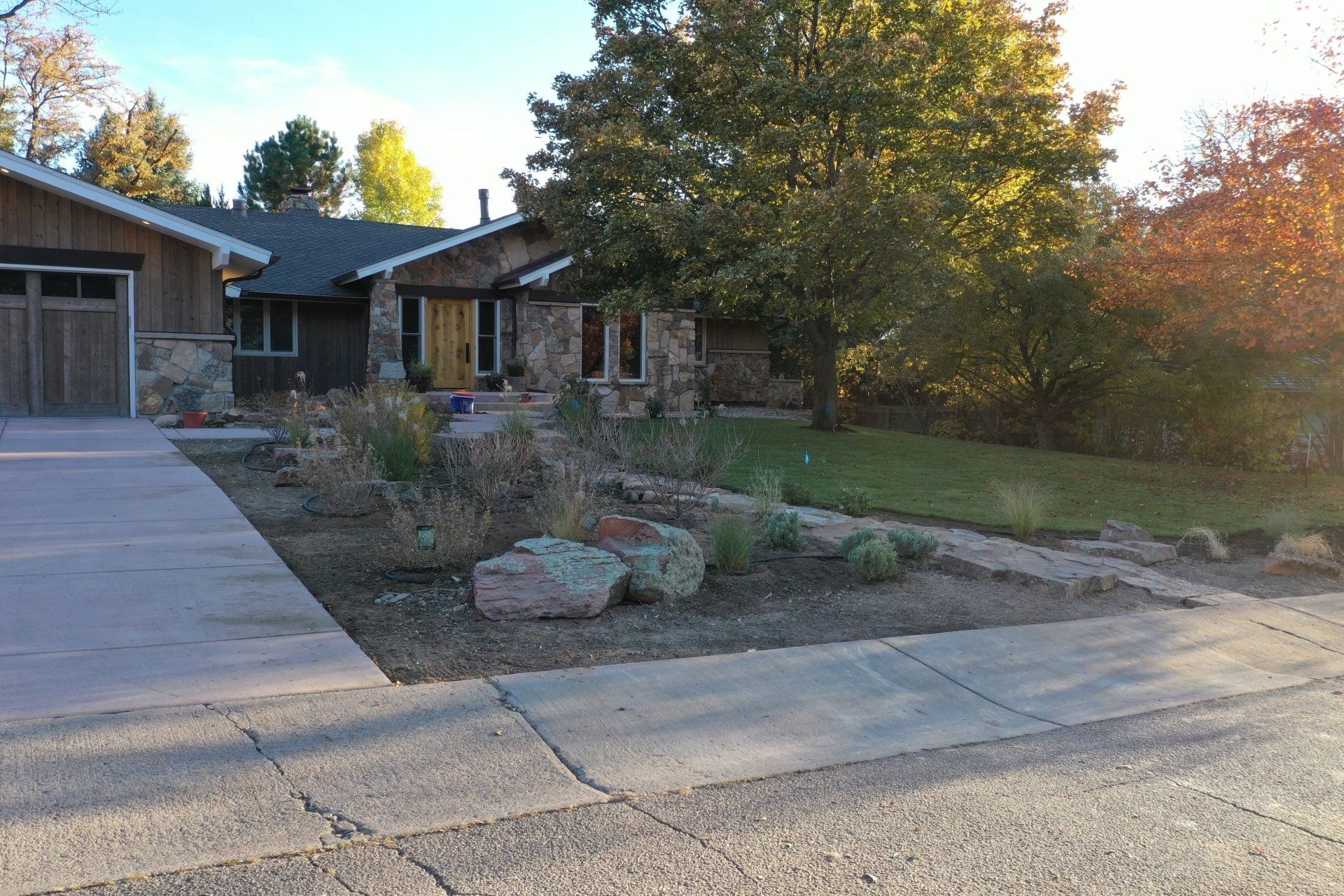 Ranch-style house with stone facade, driveway, and rock landscaping. Green grass and trees under a sunny sky.