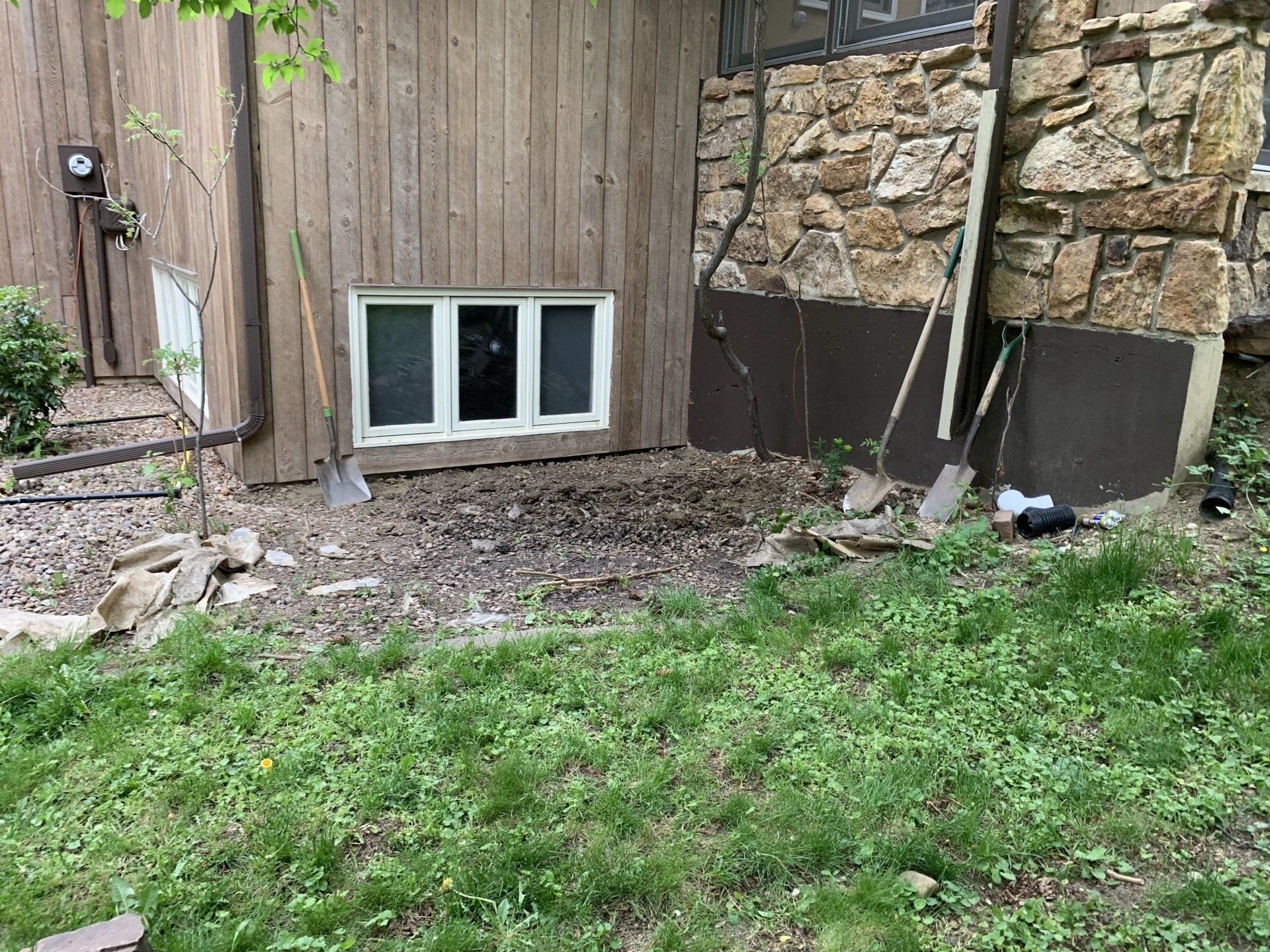 Exterior of house with dug-up dirt area near window and stone wall, tools leaning against the structure.