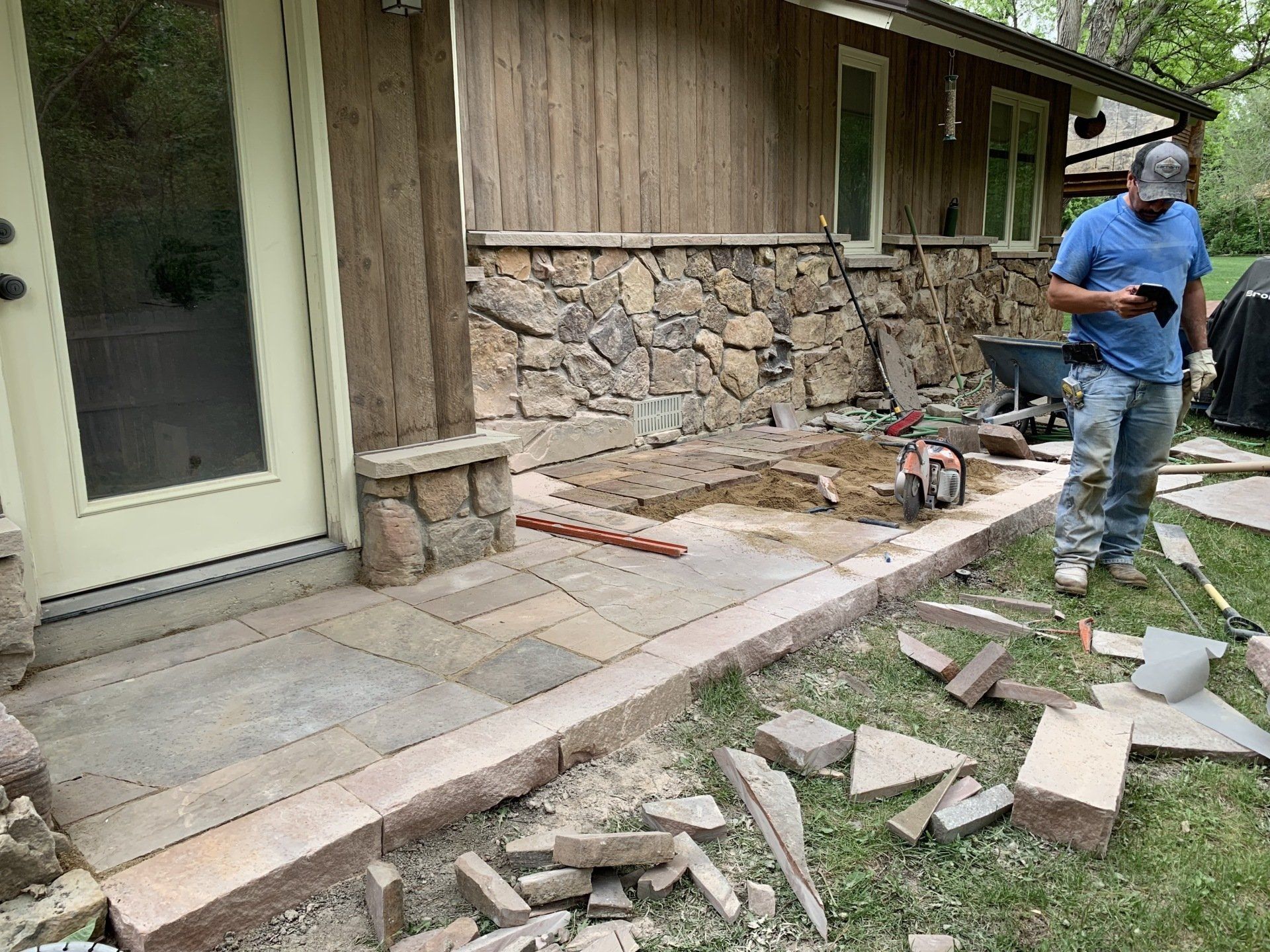 Man building a stone patio next to a house with a stone and wood exterior.