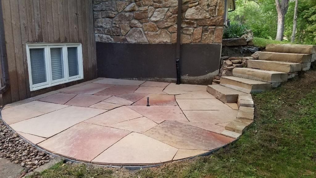 Flagstone patio with steps and stone wall, next to a wood-sided building, in a grassy setting.