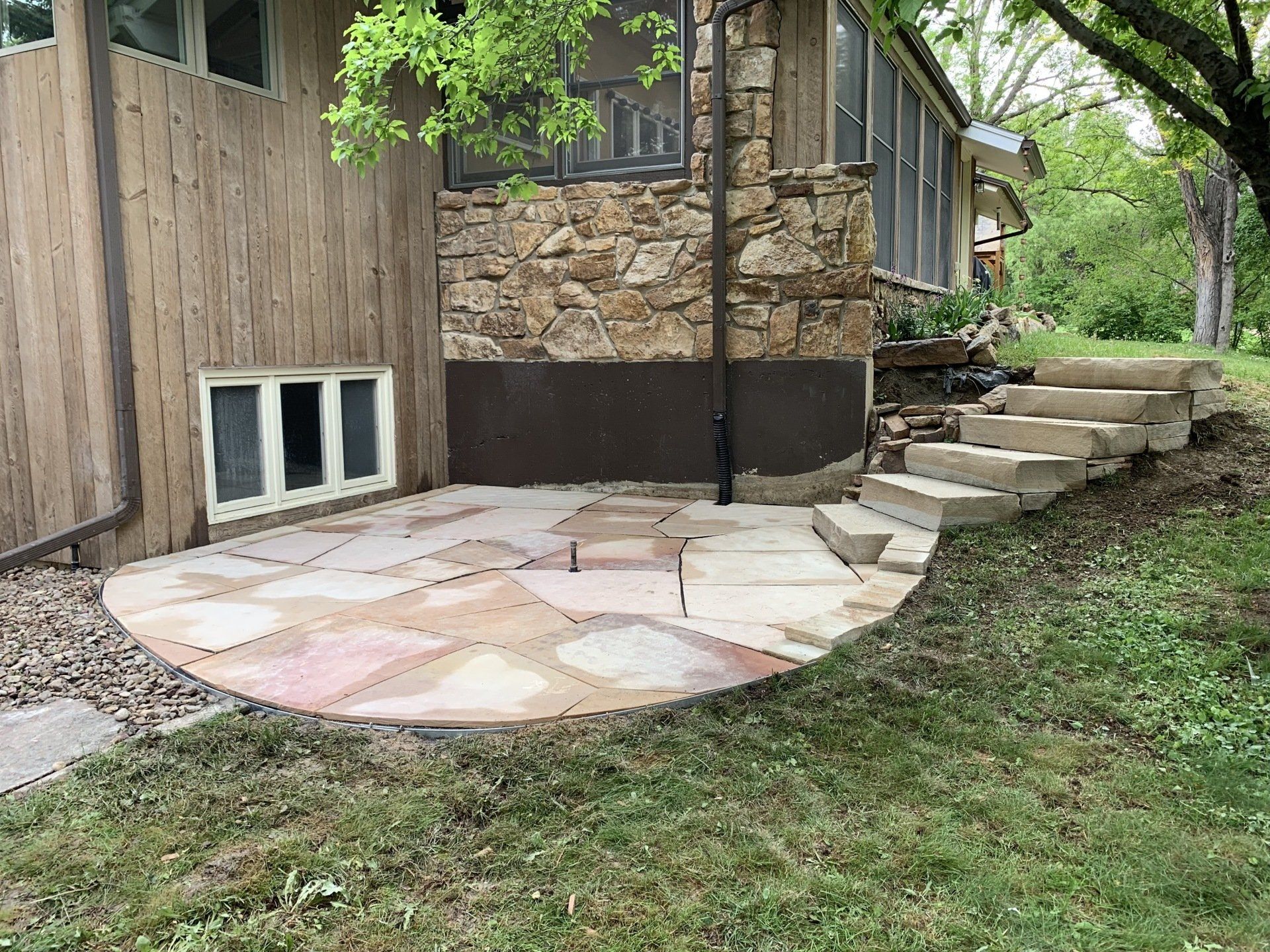 A stone patio with steps next to a house with wooden siding and a stone wall.