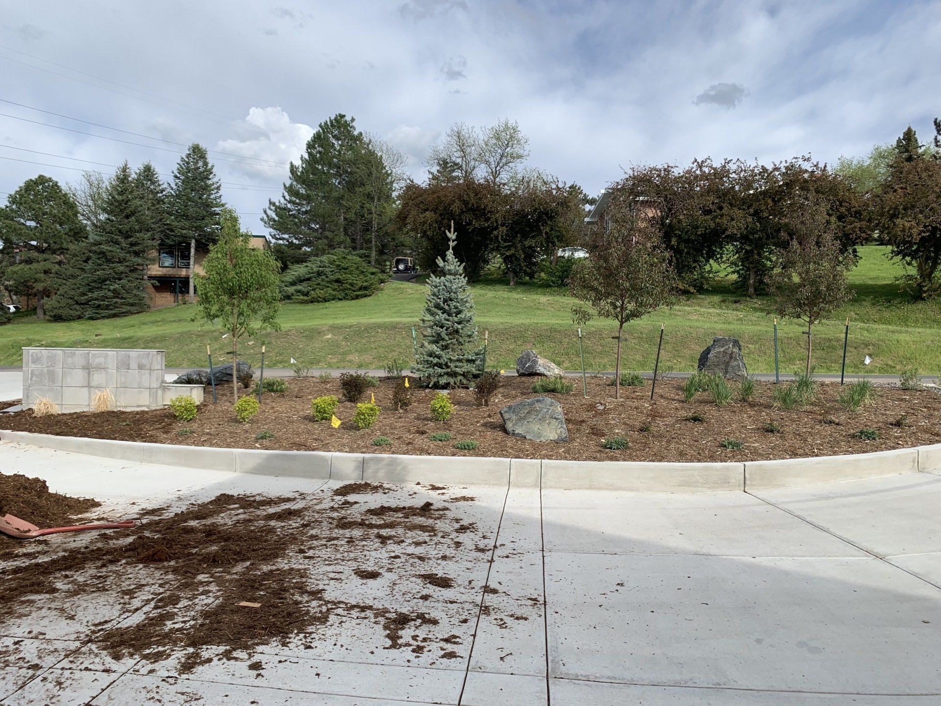 Landscaped circular island with trees, rocks, and mulch, near a concrete parking area, against a green hillside.