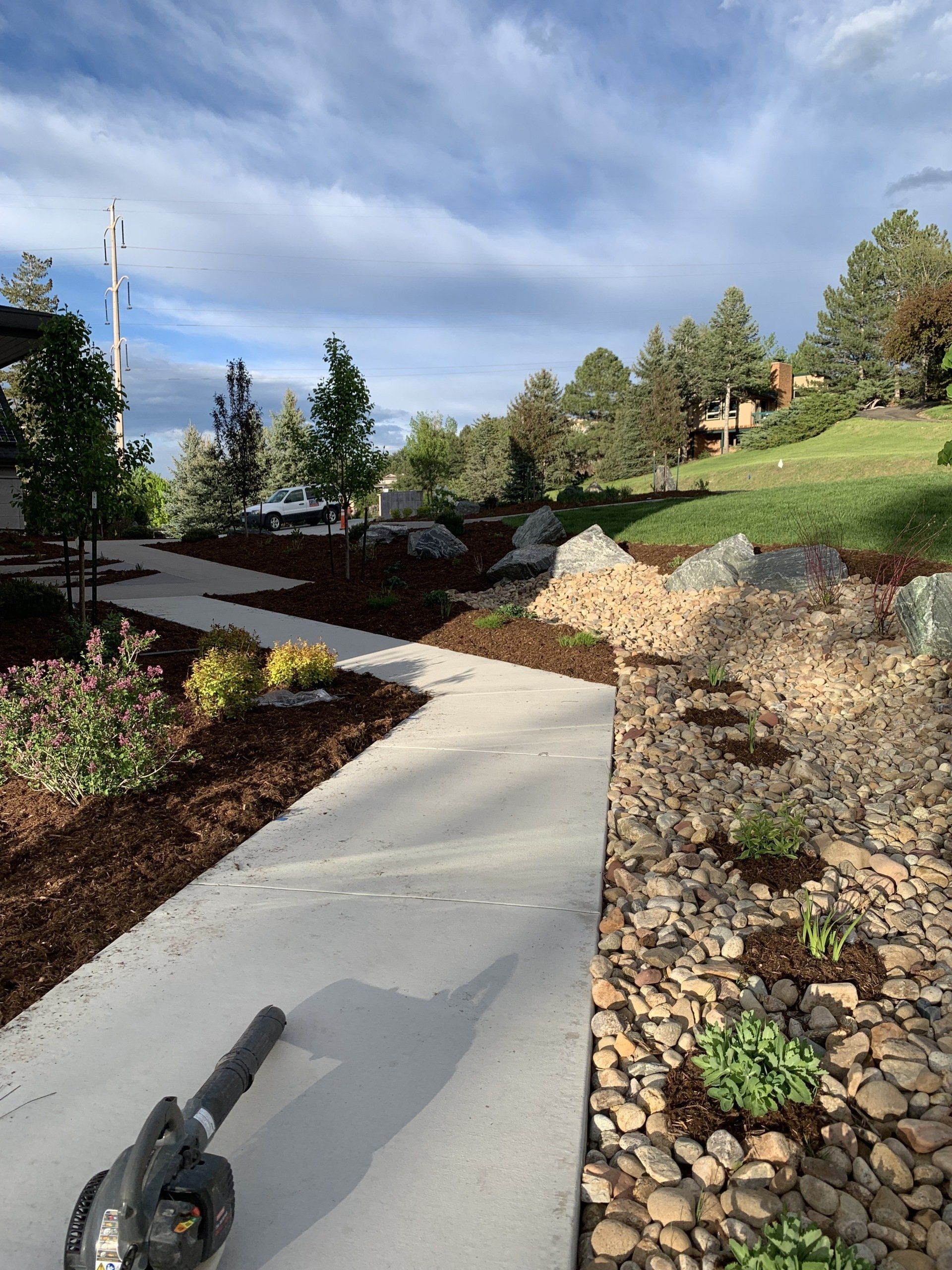 A paved walkway through a landscaped garden; a person uses a leaf blower.