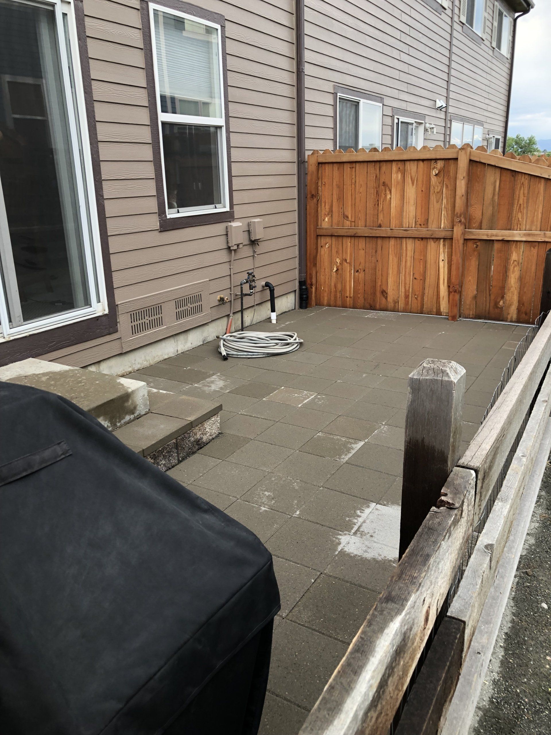 Concrete patio next to a brown wooden fence, next to a building with windows. A grill is in the foreground.