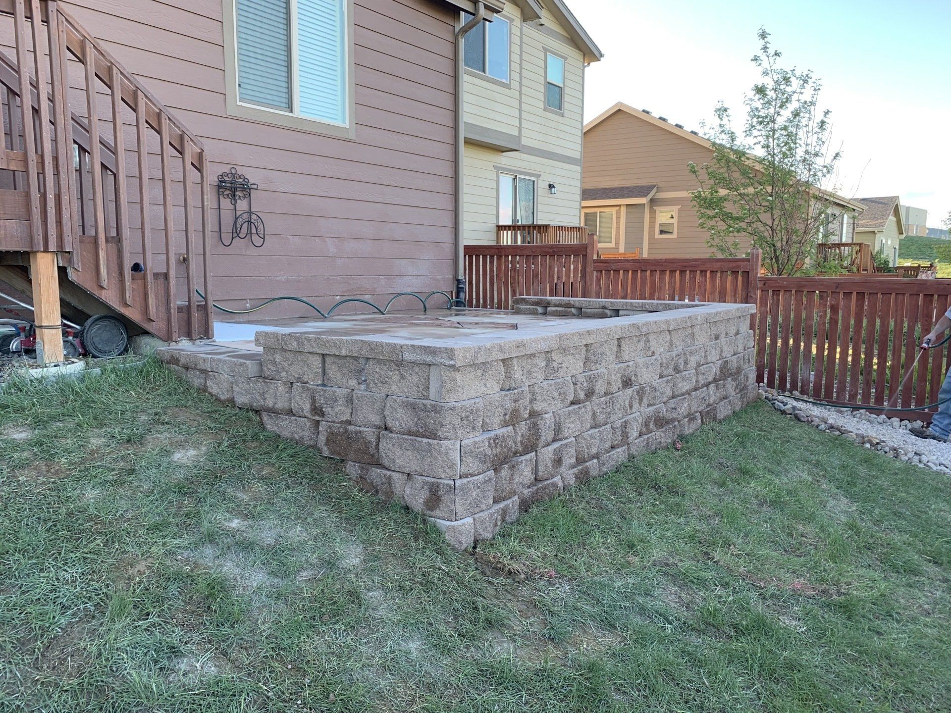 Retaining wall built of stone blocks with fire pit, next to house and wooden fence.