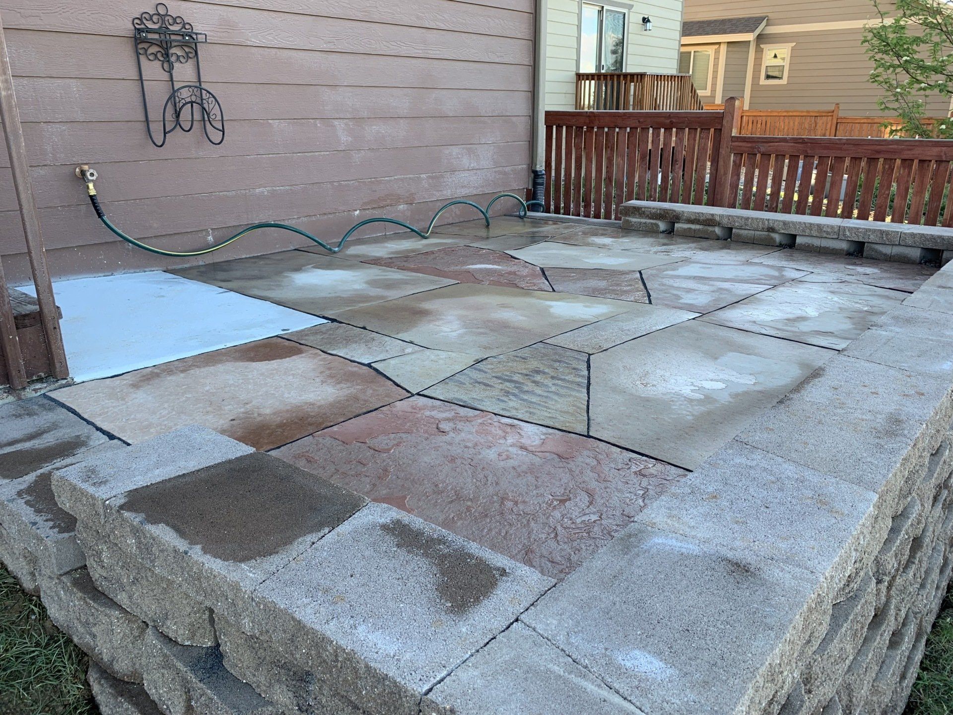 Patio with stone pavers and retaining wall; hose lying on wet surfaces, tan siding.