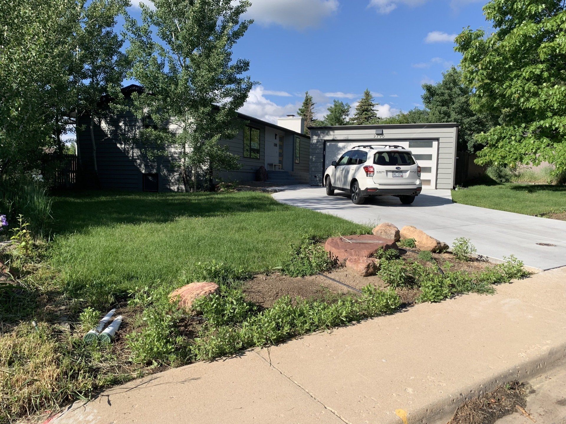 A gray house with a car in front of a garage, on a sunny day. A green lawn and sidewalk are in front.