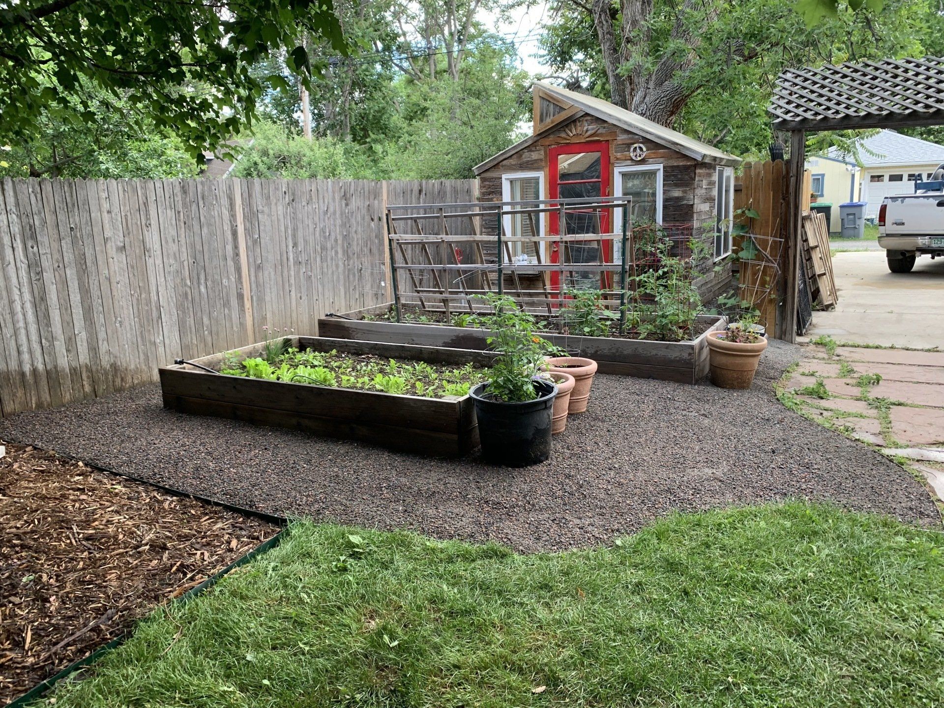 A backyard garden with raised beds, a small greenhouse, and a gravel pathway.