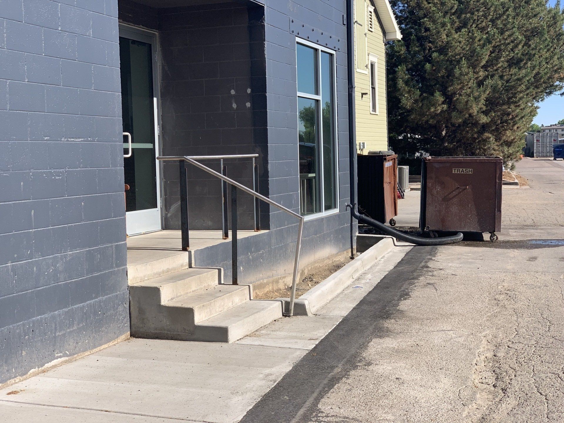Gray building entrance with steps, handrail, and ramp, next to a dumpster on concrete.