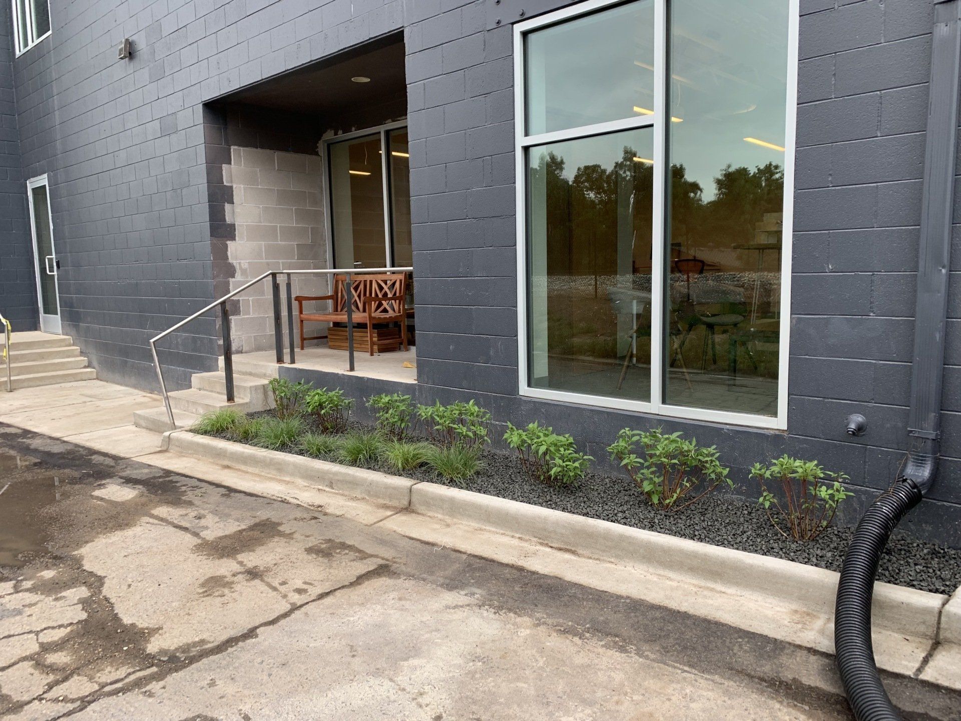 Exterior of a dark gray building with steps, a window, landscaping, and a bench visible inside.