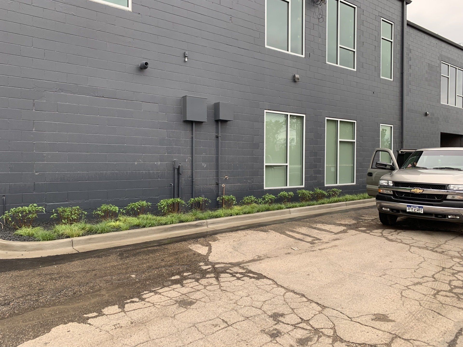 A dark gray building with windows and landscaping next to a gray pickup truck parked on cracked asphalt.