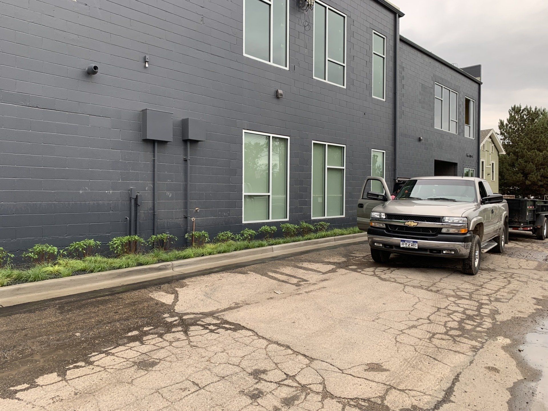 A silver pickup truck parked next to a dark gray building. The door is open, and the pavement is cracked.
