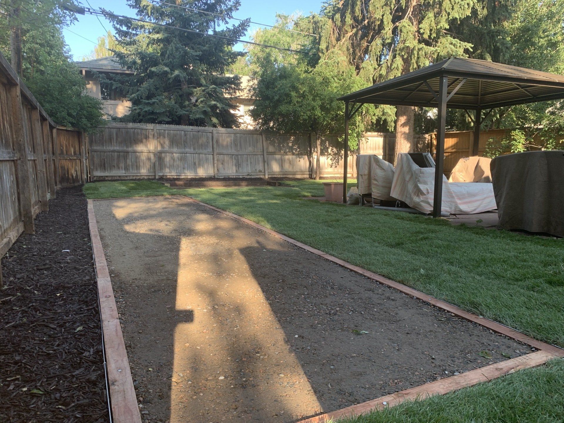 Backyard with a gravel area bordered by wood, and a gazebo on the right, surrounded by green grass and a wooden fence.