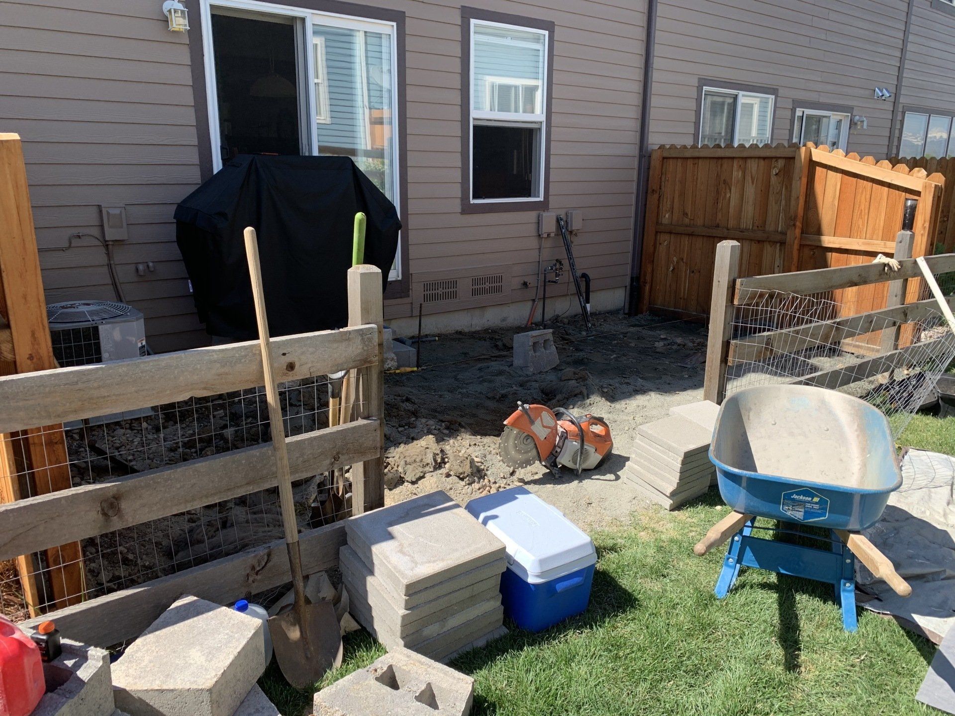 Backyard patio project: Blocks, tools, and wheelbarrow on grass near a fence and house.