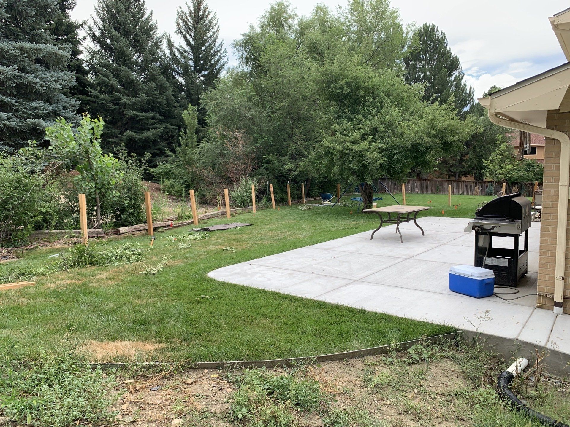Lawn with patio, grill, and fence posts. Trees and a house are in the background.