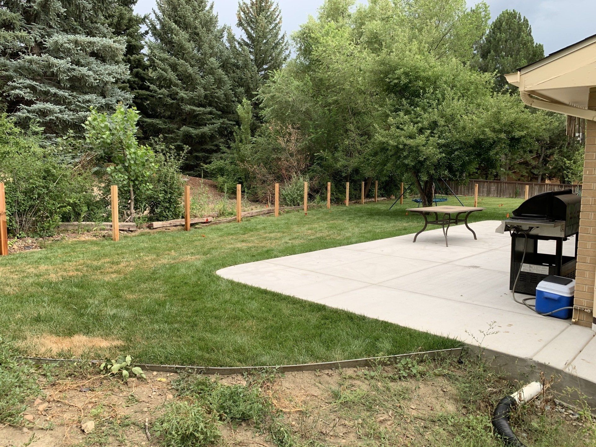 A backyard with a patio, grill, and fence posts; a table and lawn with trees in the background.