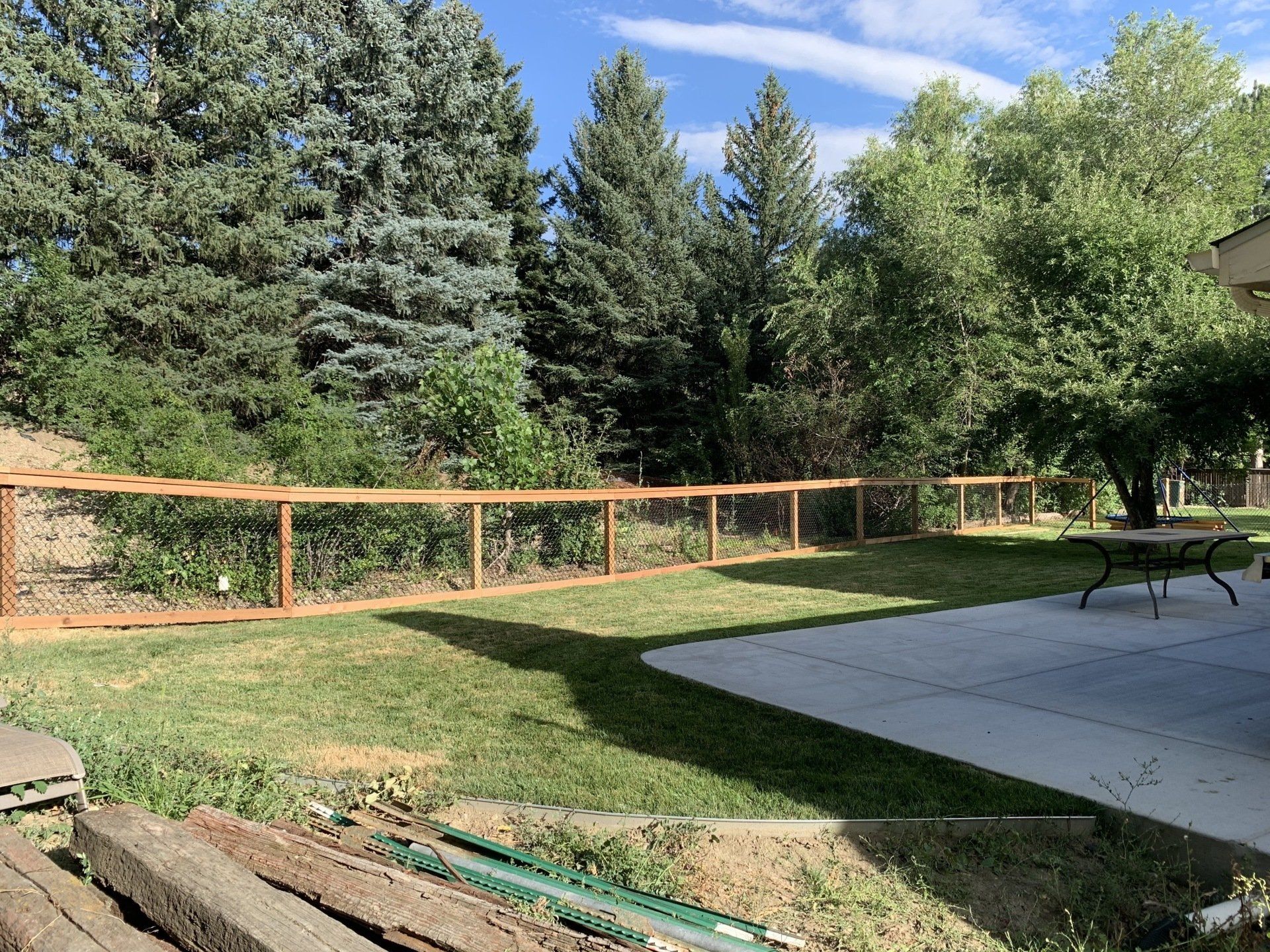 Wooden fence enclosing a backyard with green grass, trees, and a concrete patio.
