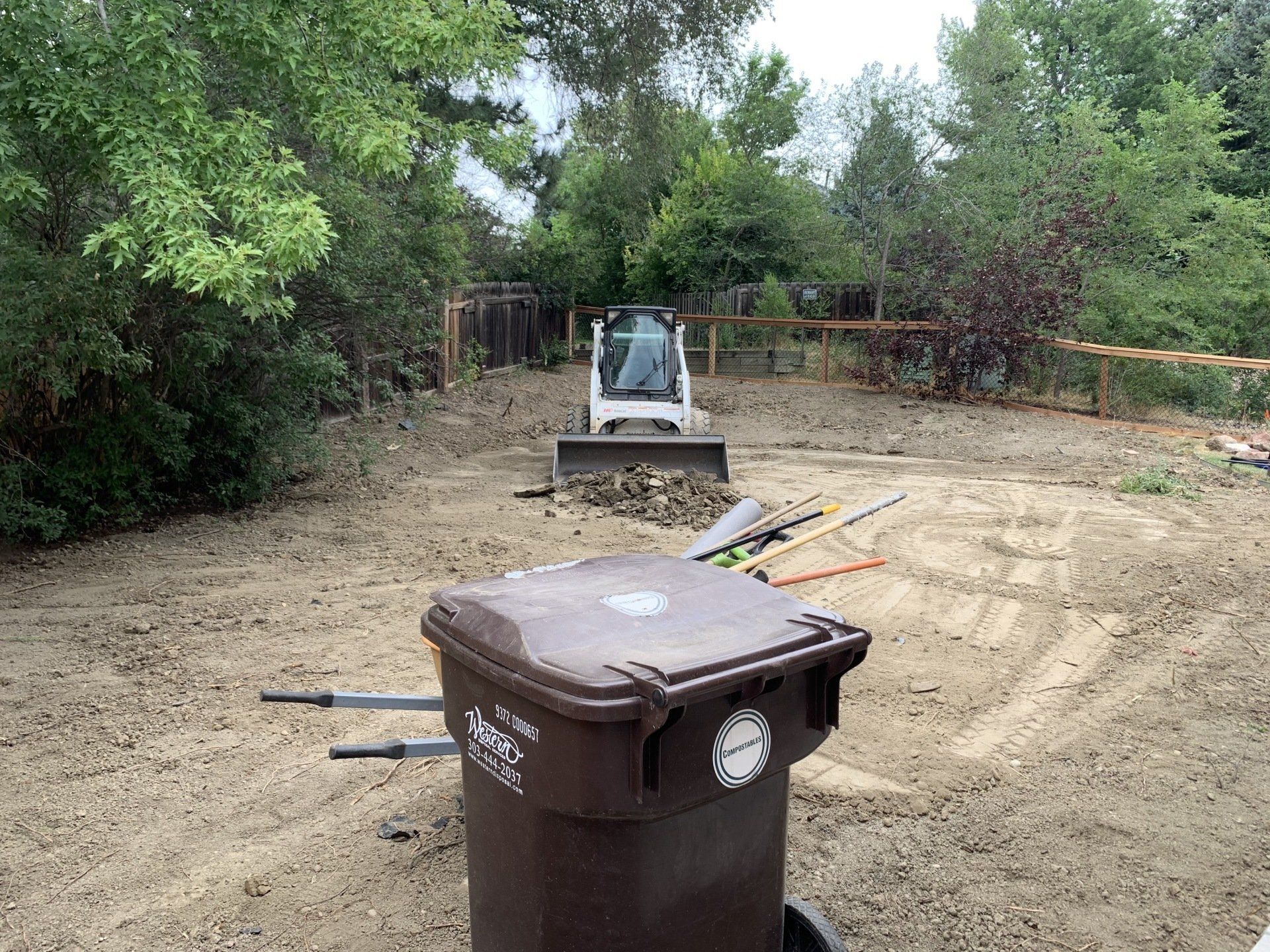 Brown trash bin in foreground, small bulldozer moving dirt in backyard. Trees, fence in background.