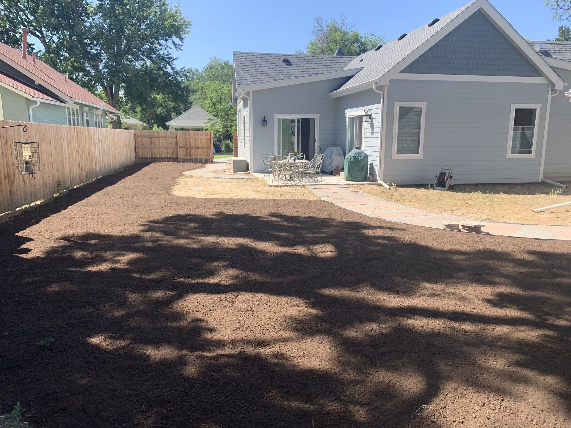 Backyard with freshly tilled soil, a light blue house, wooden fence, and trees casting shadows.
