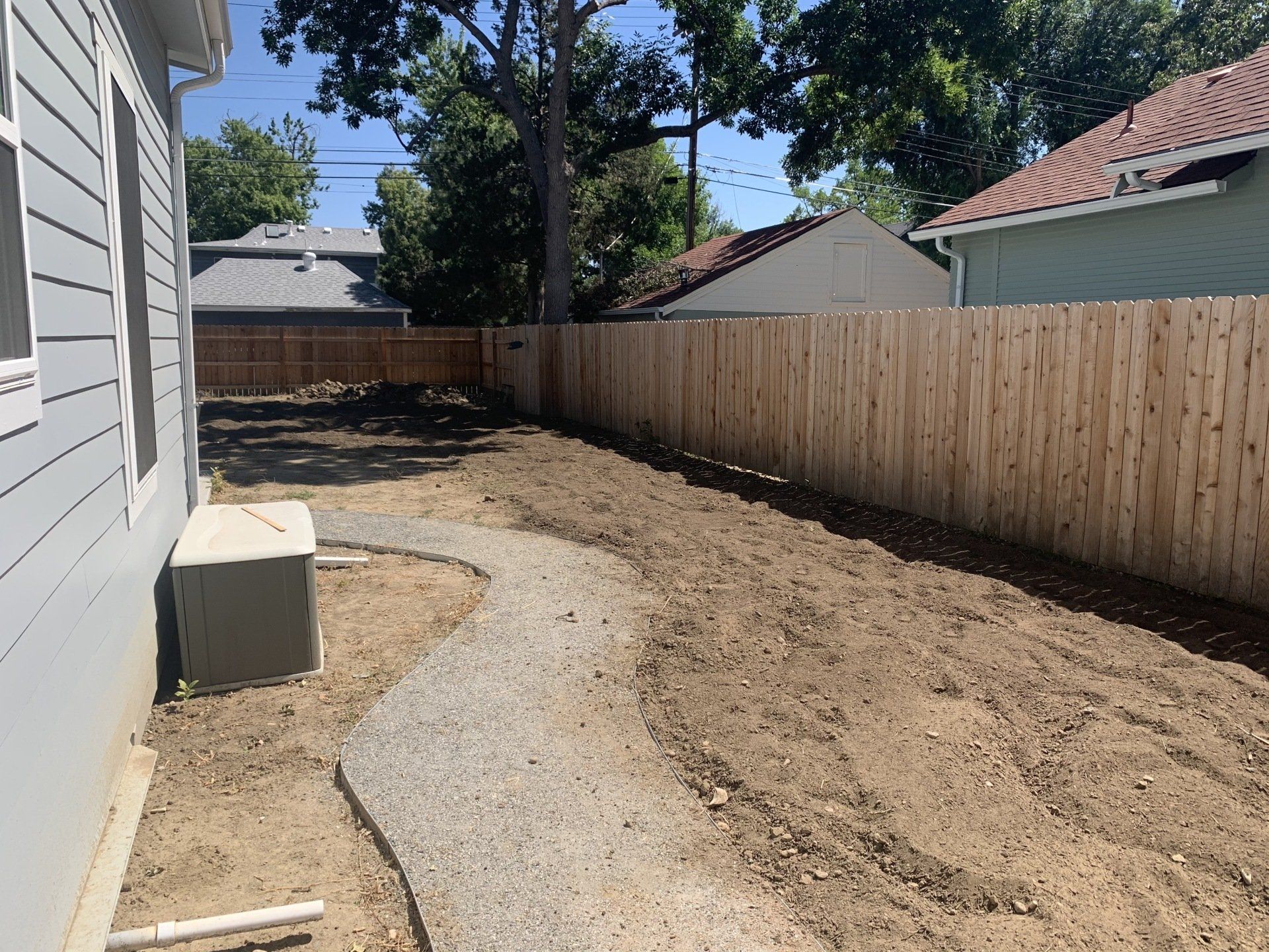 Backyard with gravel path, wooden fence, and freshly tilled soil. Gray house on the left.
