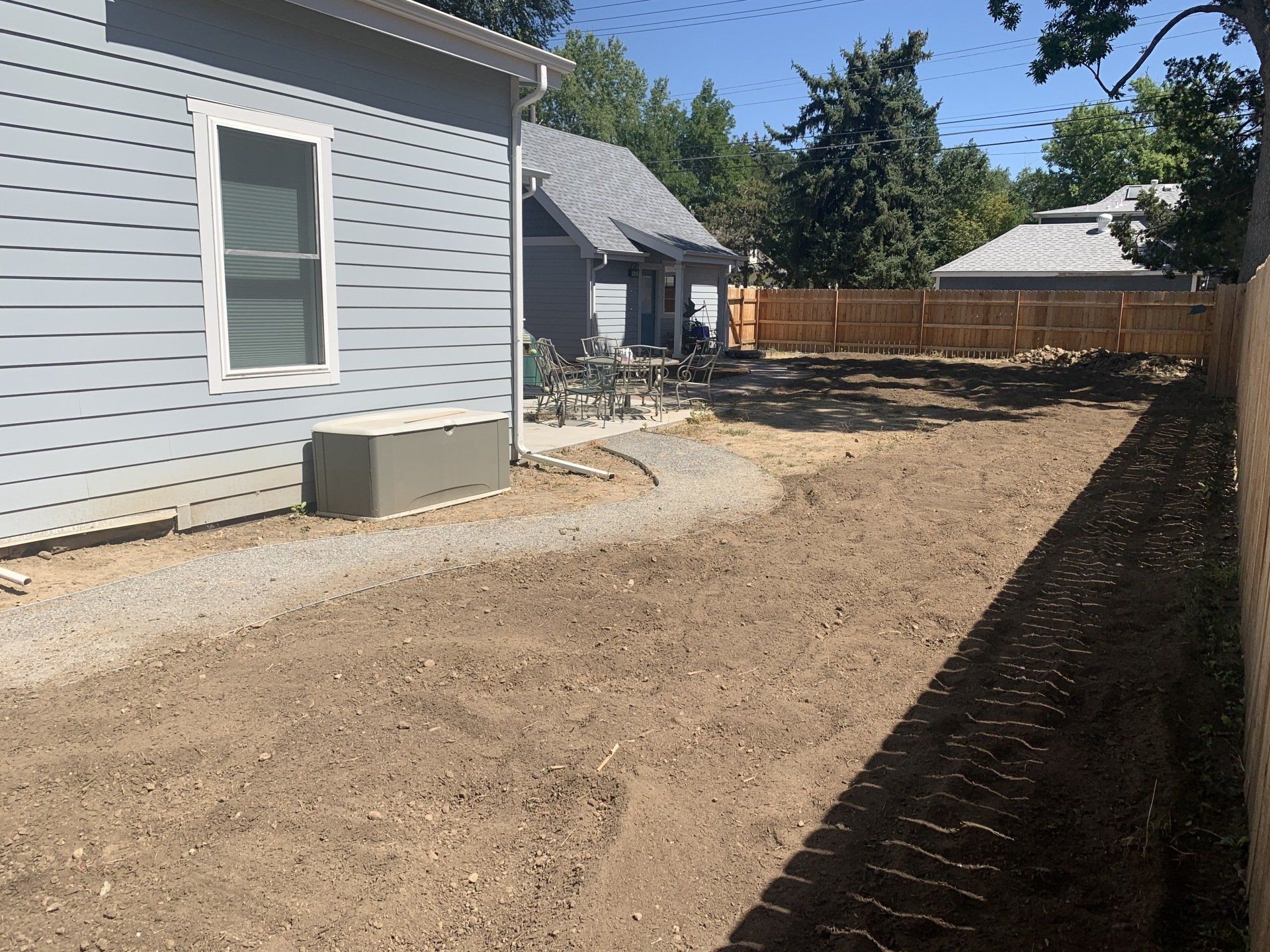 Side yard with a gravel path, dirt, and fences; a light blue house is on the left.