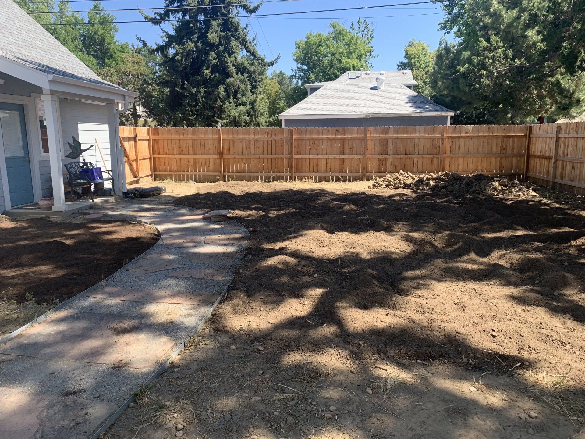 A backyard with a wooden fence and dirt path. Newly tilled soil in the yard, sunny day.