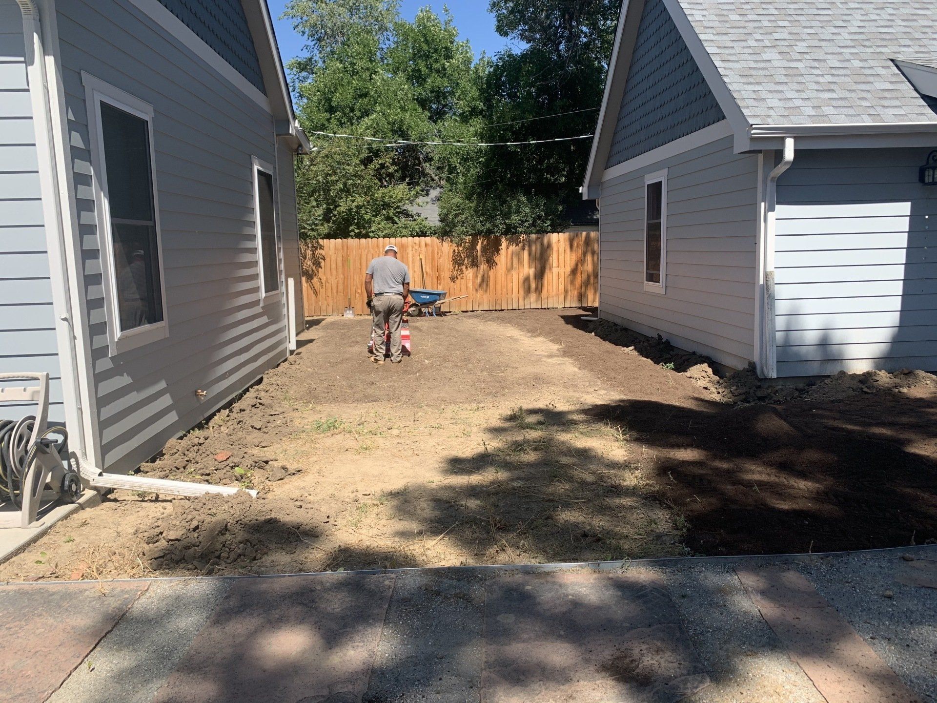 Man tilling soil between two houses, preparing for landscaping. Brown dirt, wooden fence, sunny day.