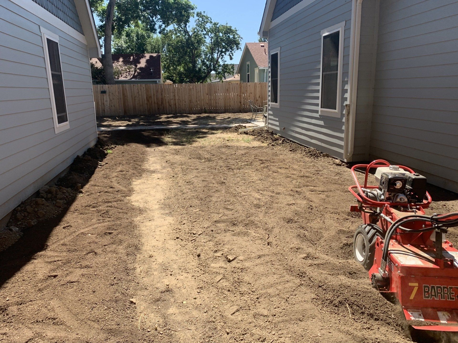 Backyard space with dirt, between two gray houses. A small red machine is on the right side, a wooden fence in the back.