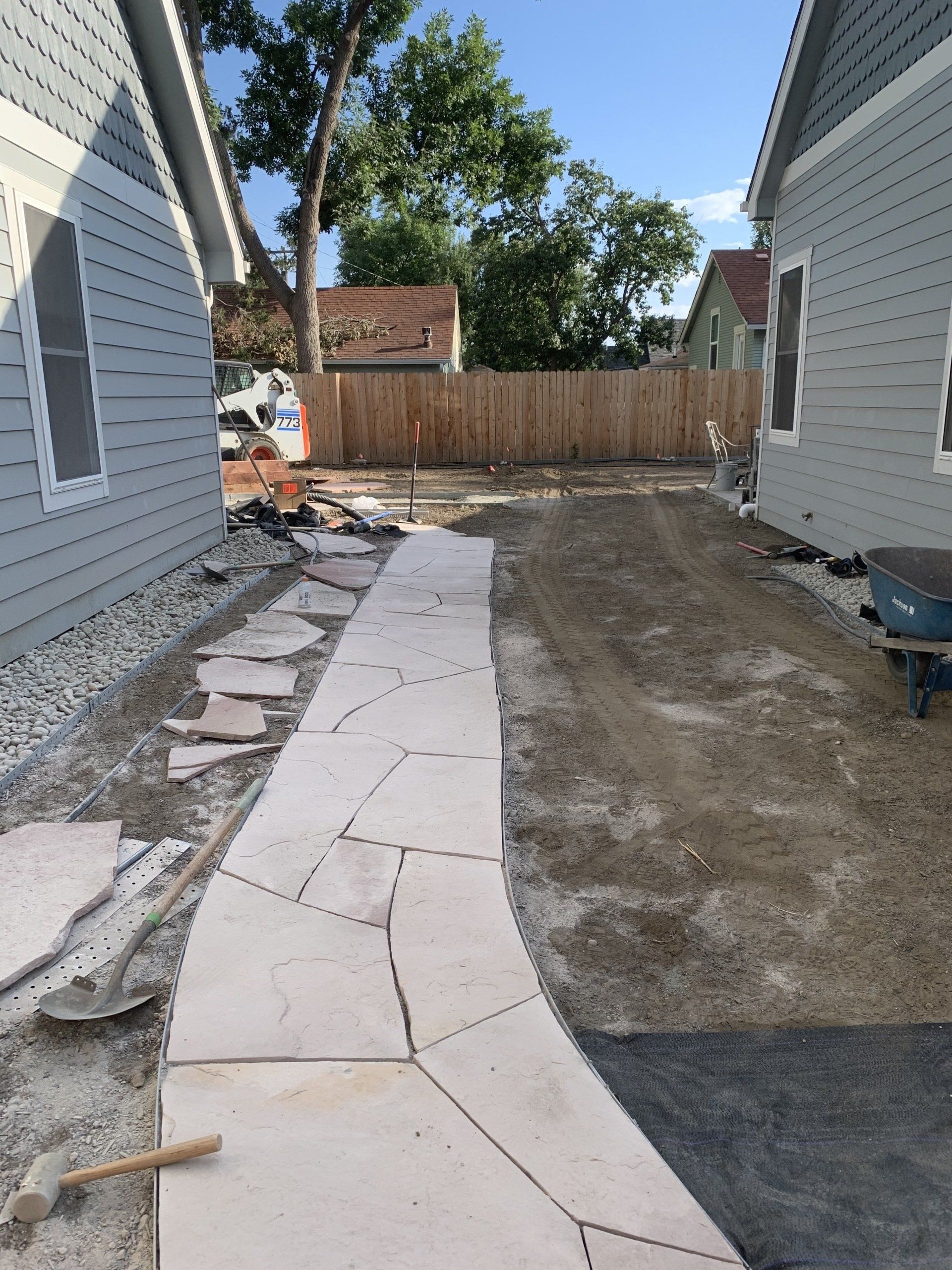 Stone pathway construction between two gray houses, leading to a wooden fence.