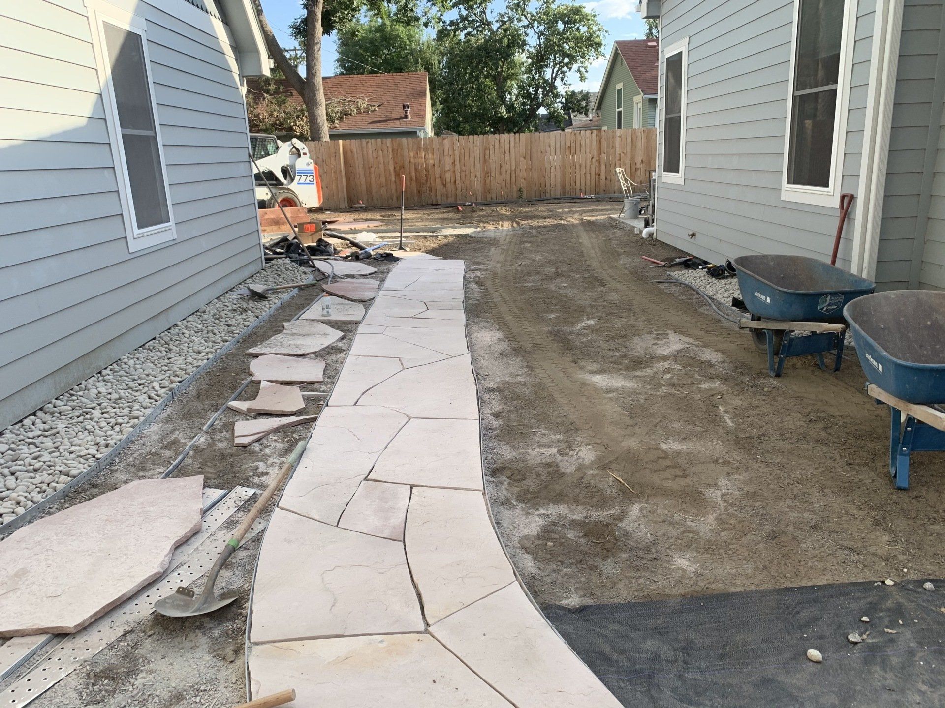 A stone pathway being constructed between two houses, with gravel and dirt visible.