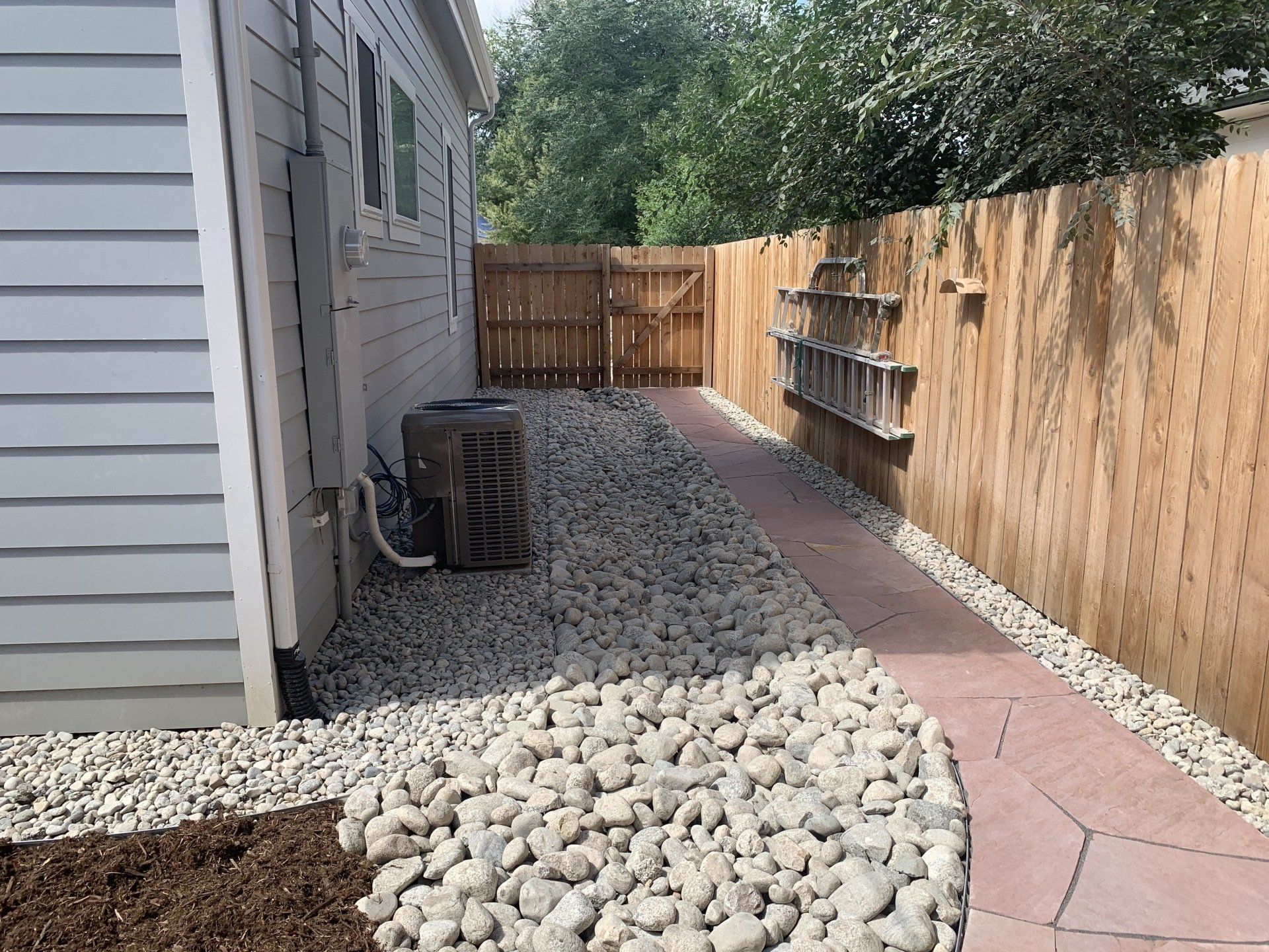 Gravel and red-paver path alongside a wooden fence and the side of a house, leading to a gate.