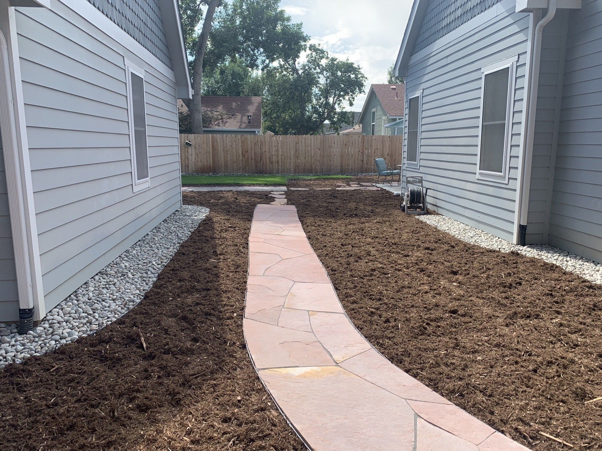 Pathway between two gray houses with a wooden fence in the background.
