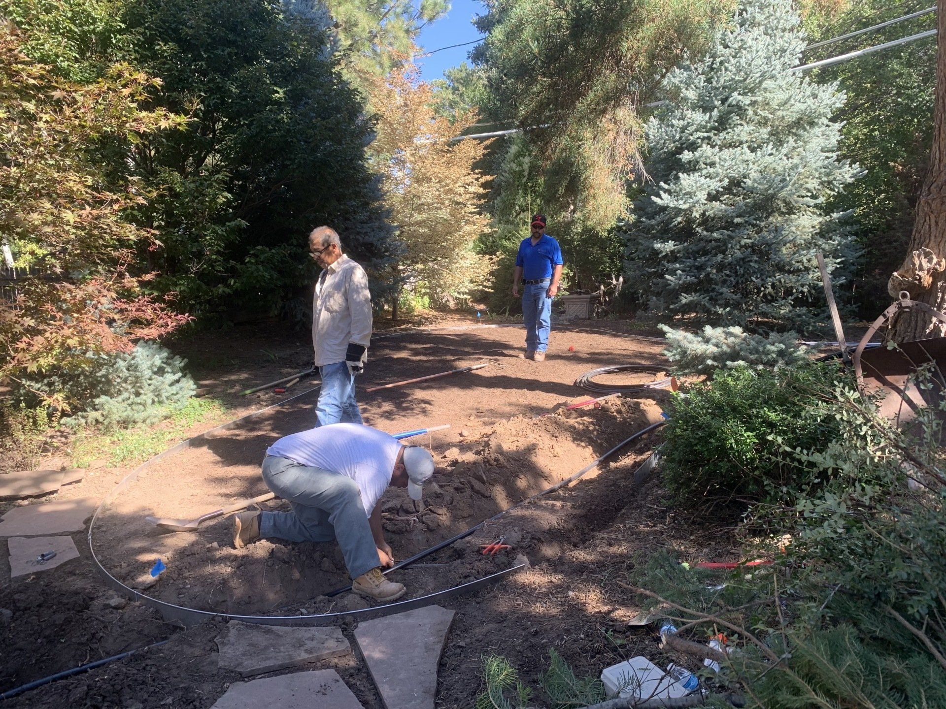 Three men working in a garden bed. One kneels, two stand. Trees and blue sky background.