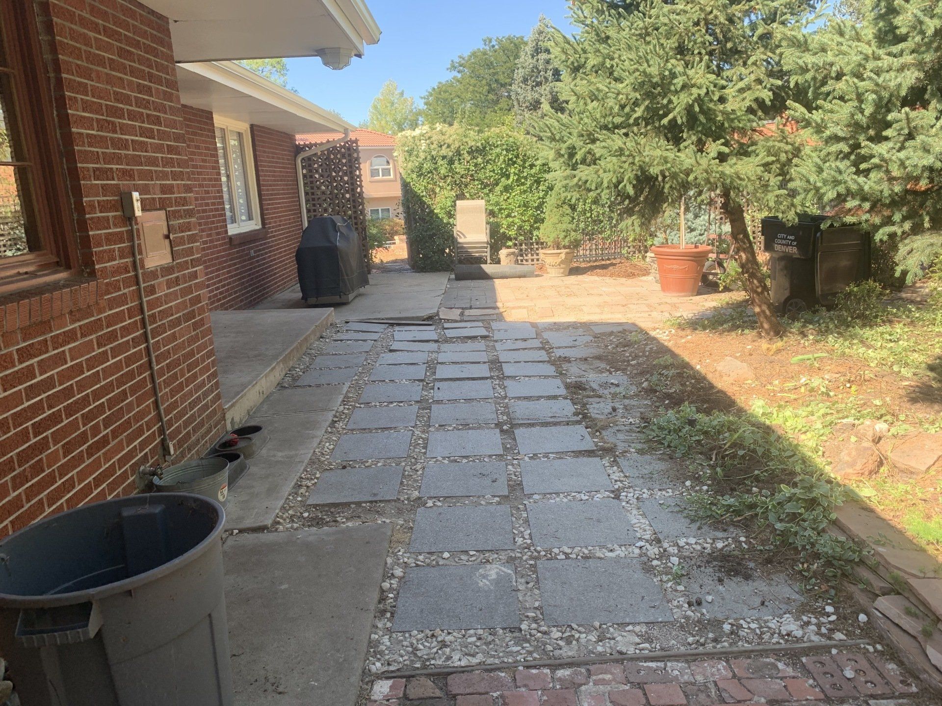 A brick house backyard with a stone walkway, greenery, a trash can, and a grill.