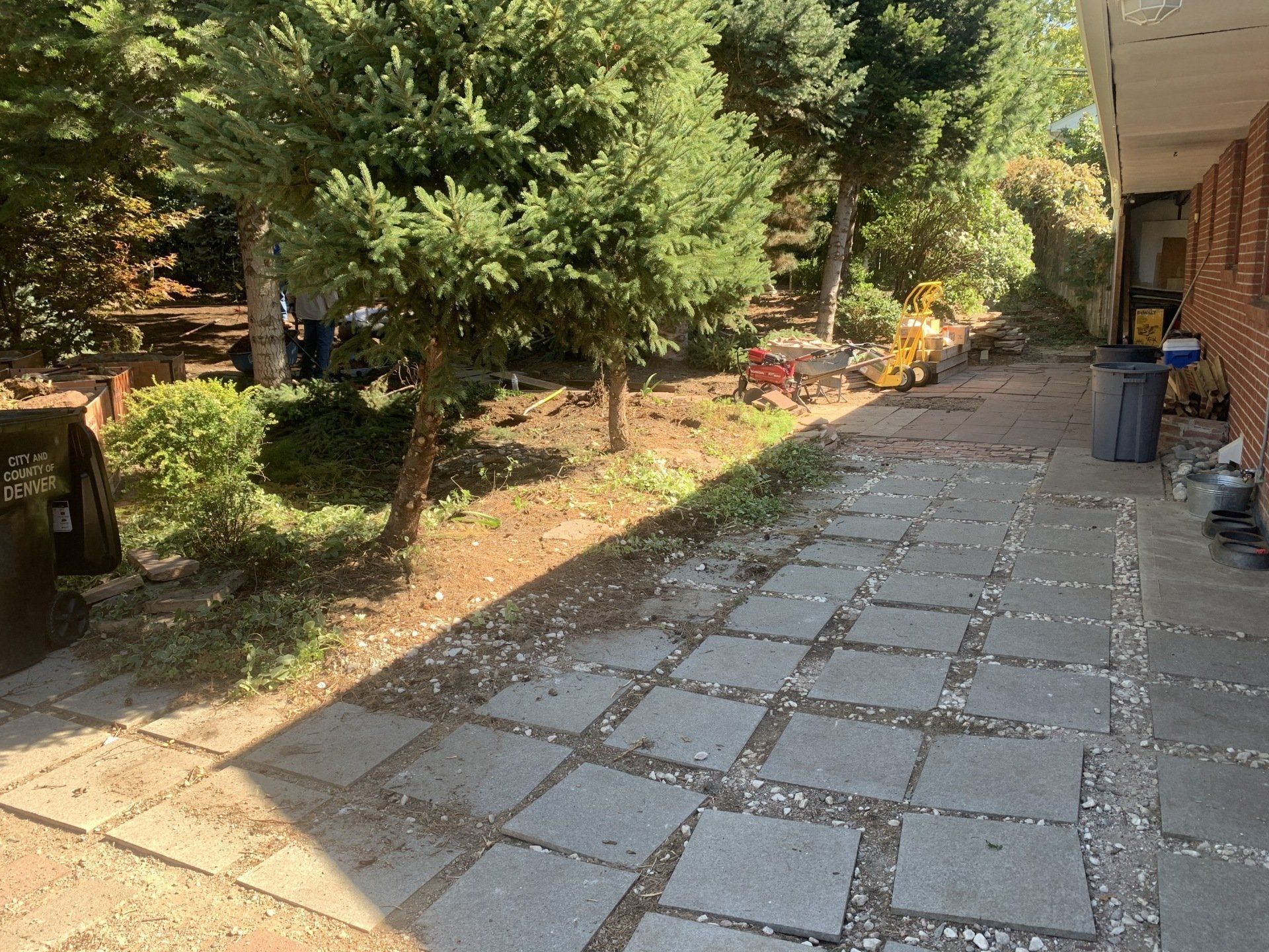 A paved walkway alongside a building and trees; construction equipment and debris are visible.