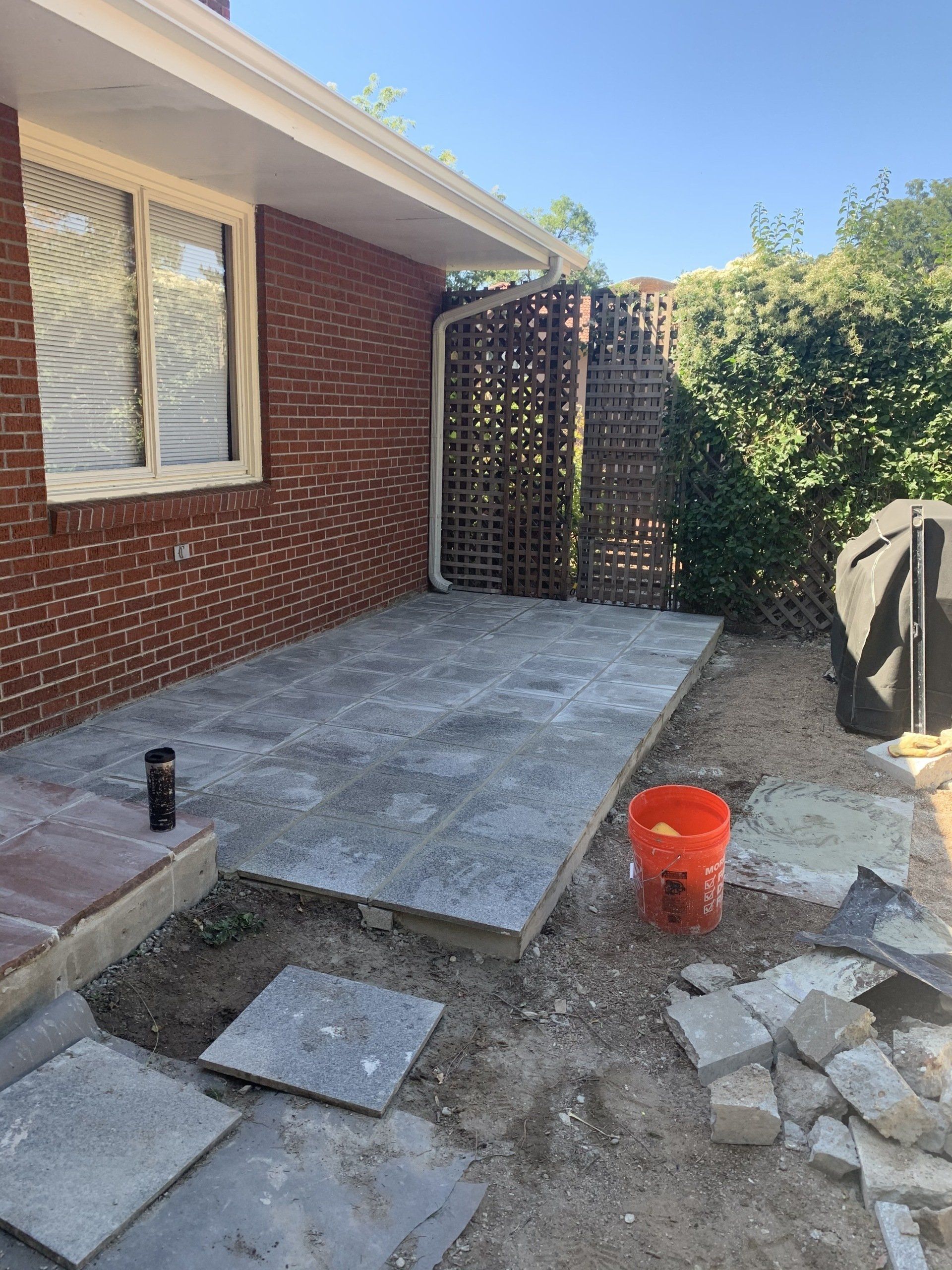 Patio with gray pavers being installed. Brick wall, lattice fence, orange bucket, and loose pavers are visible.