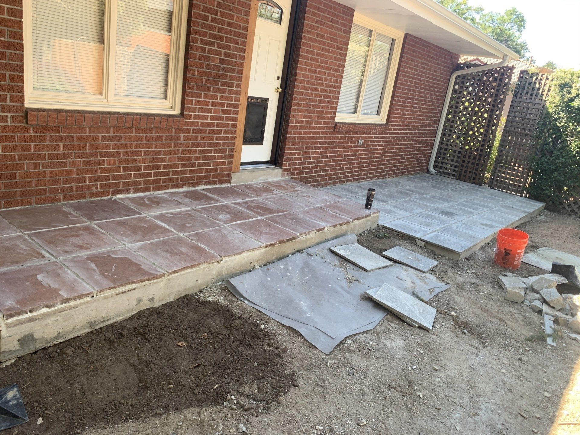 Front porch under construction: red brick building, concrete slabs, stone pavers, dirt, orange bucket.