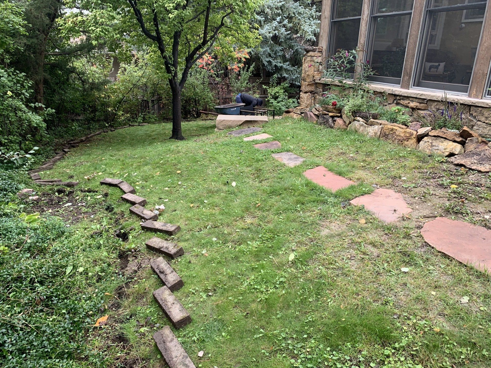 Lawn with stepping stones leading to a tree. Stone building on the right, surrounded by greenery.