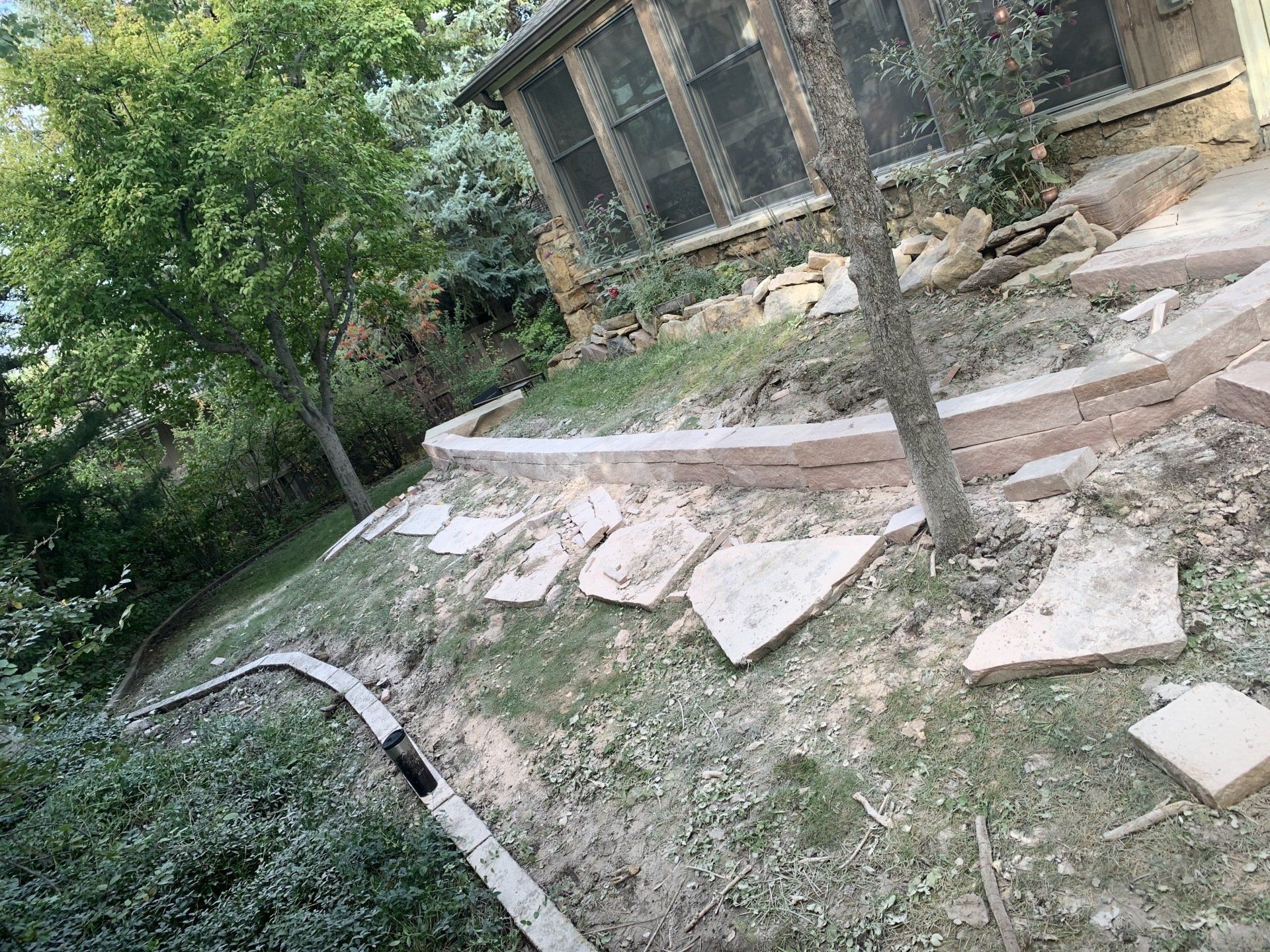 Stone walkway construction with retaining wall near a house, on a hillside.