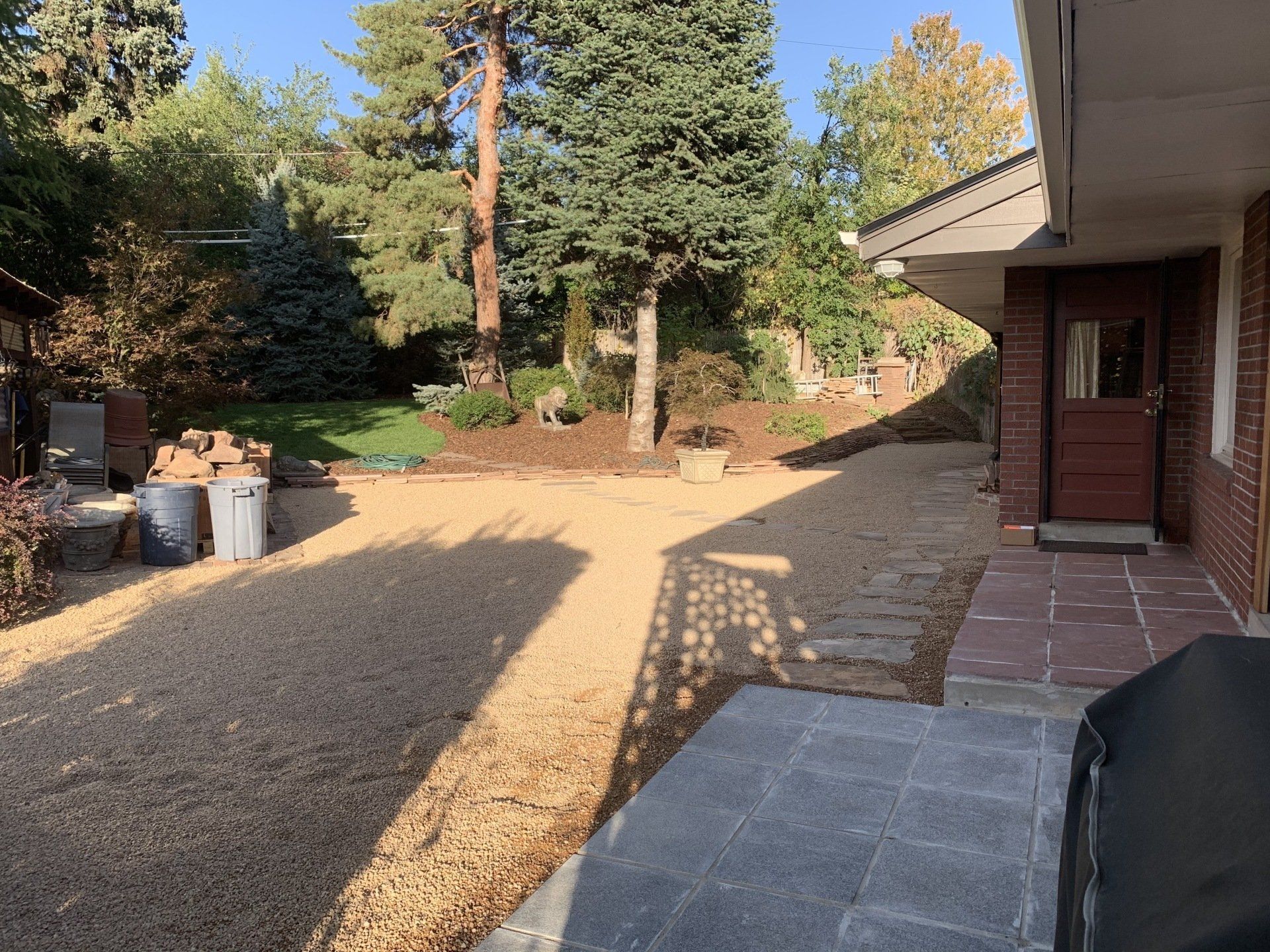 Gravel yard with trees, building, woodpile, and shadow patterns from sunlight.