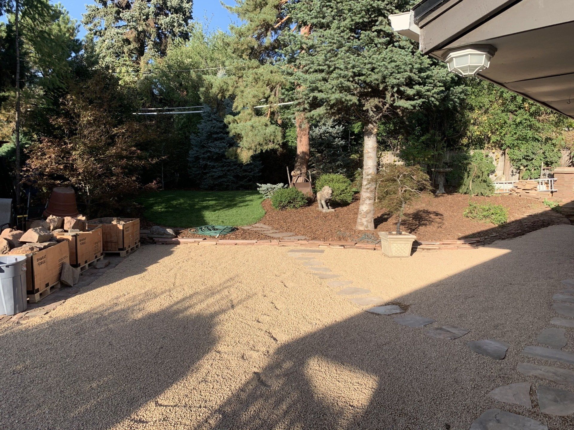 Gravel driveway with materials, leading to a landscaped backyard with trees, grass, and a sunny sky.