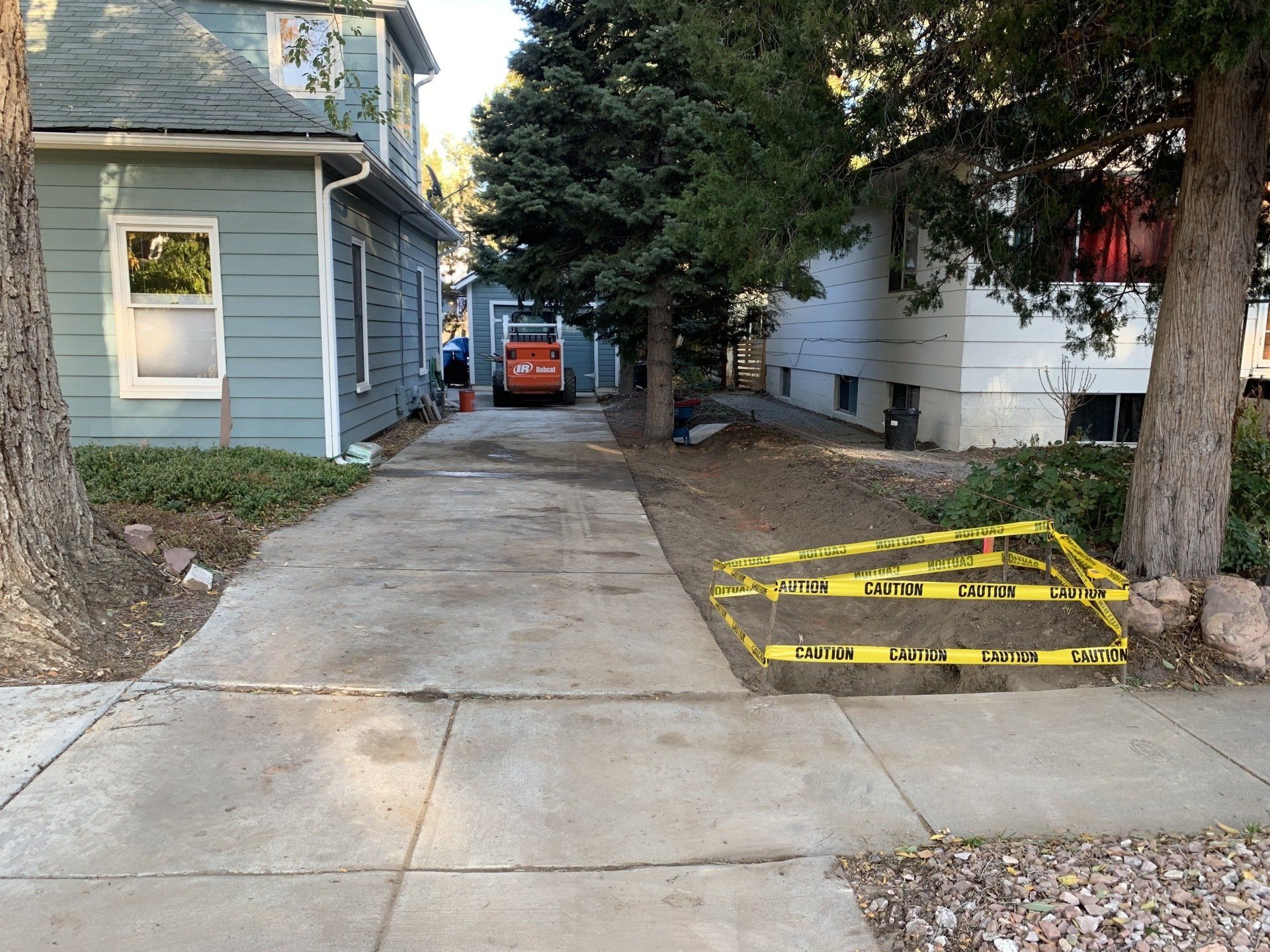Concrete driveway with caution tape, flanked by buildings and trees. Small excavator visible.