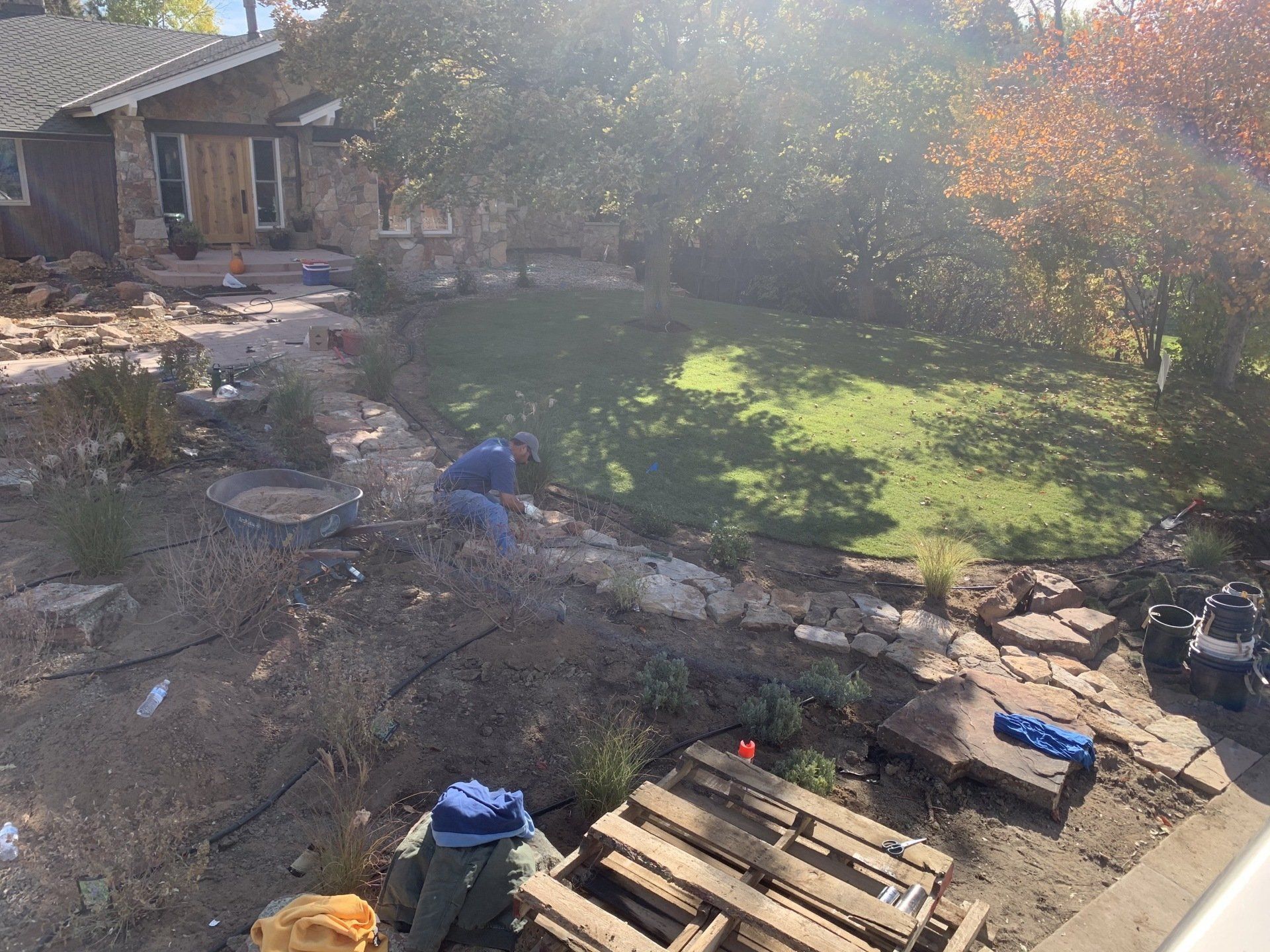 Construction site: workers near house with exposed stonework, dirt, pallets, and a small lawn.