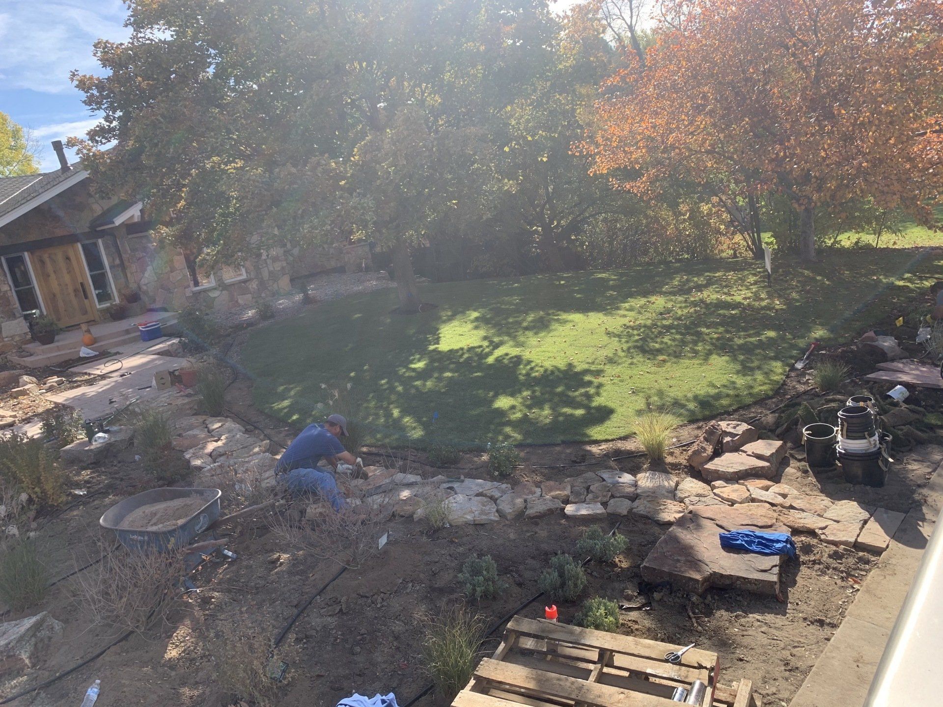 Man landscaping backyard with stone wall and lawn surrounded by trees and a house.
